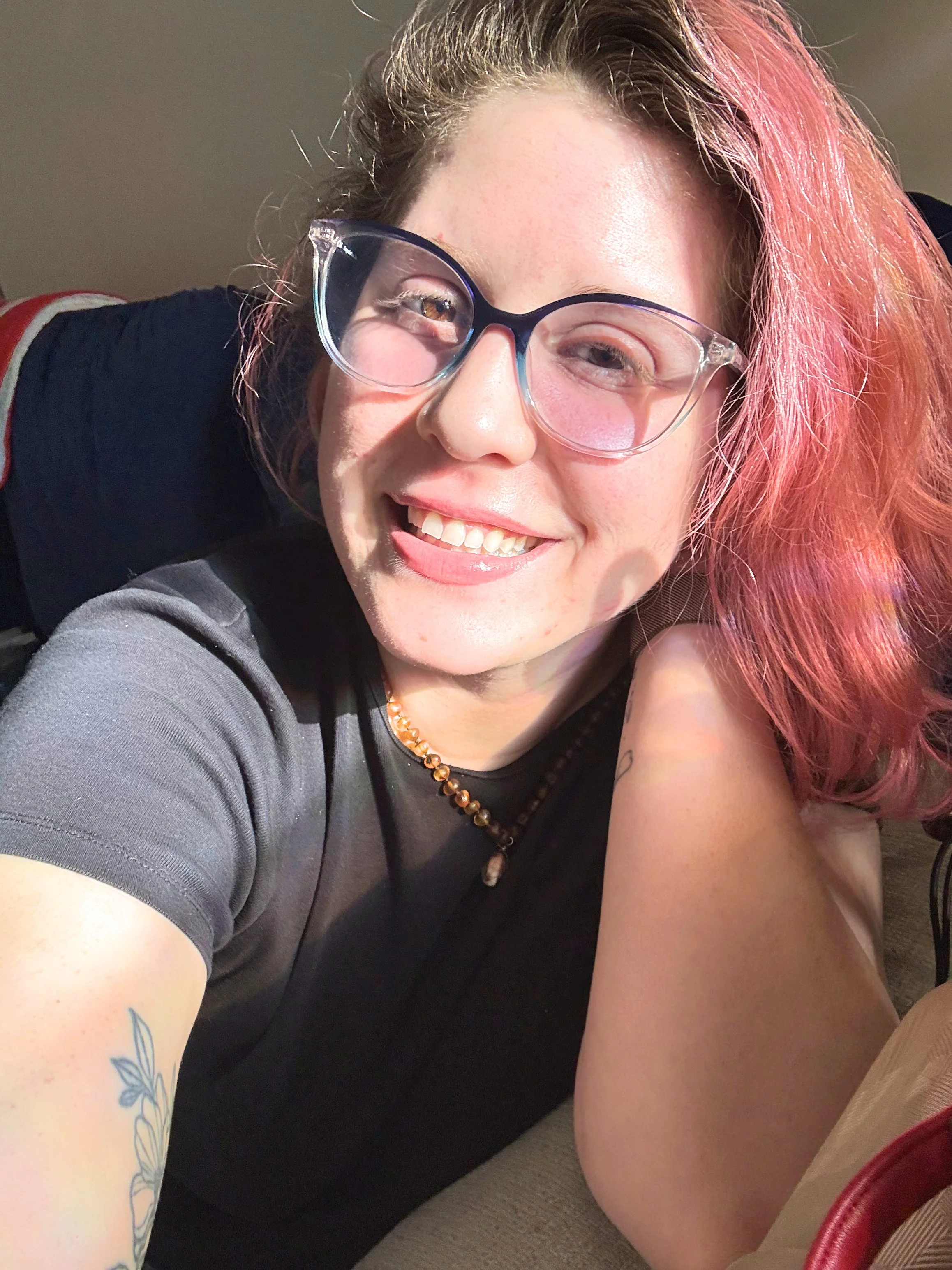 A smiling woman with pink hair, wearing glasses and a black shirt, sitting indoors with her arm resting on her knee, showing a tattoo of a flower on her forearm.