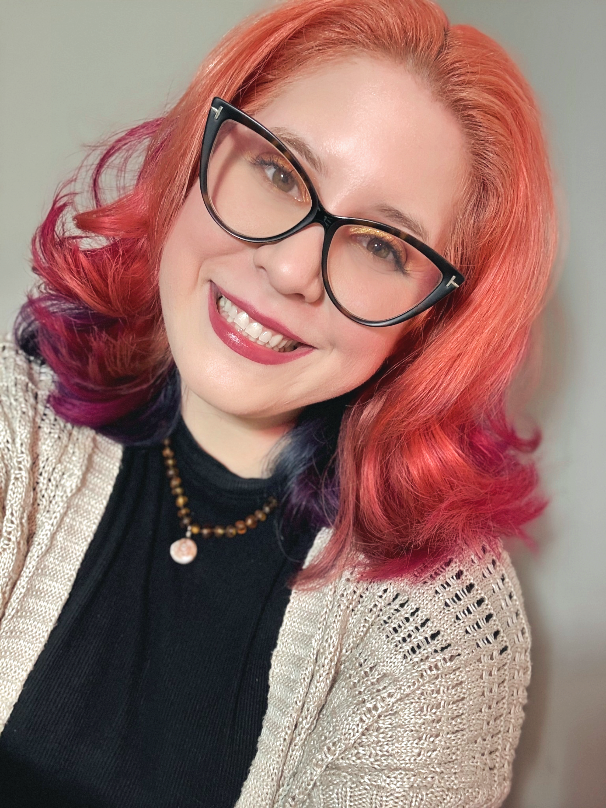 A woman with orange wavy hair wearing black glasses, a beige knitted cardigan, a black top, and a necklace, smiling at the camera.