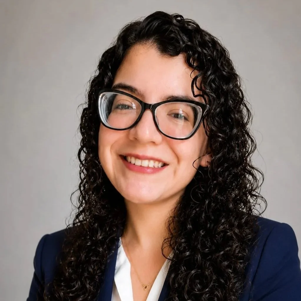 Mujer con cabello rizado y gafas grandes, sonriendo, con ropa formal, fondo gris.