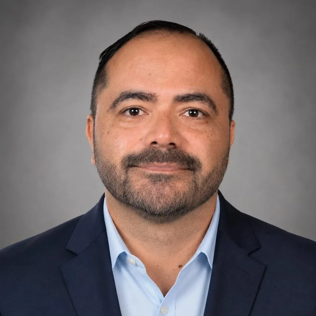 Retrato de un hombre con barba y cabello oscuro, vestido con saco azul y camisa celeste, con fondo gris.