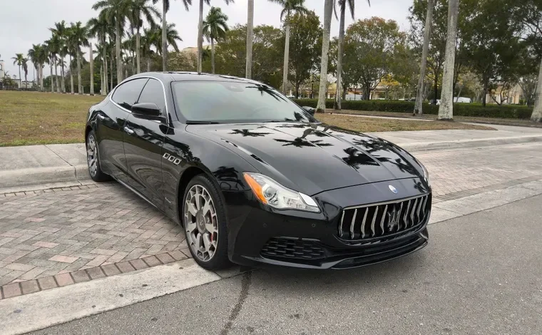 Black Maserati luxury sedan parked on a brick driveway with palm trees and greenery in the background.