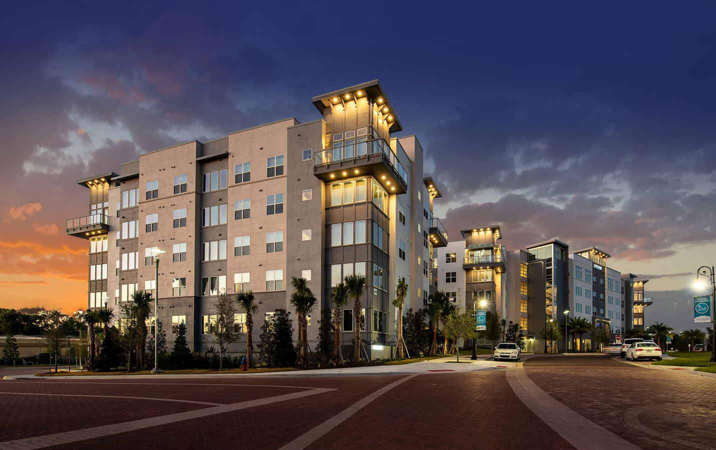 Modern apartment buildings at sunset with lit balconies, palm trees, streetlights, and parked cars.