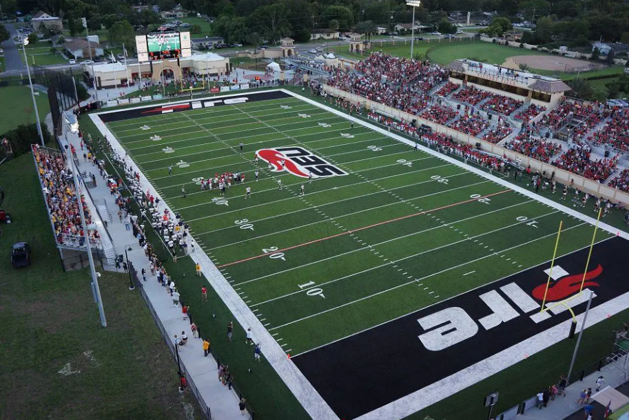 Aerial view of a football stadium with fans seated in stands and players on the field during a game, featuring a large logo at the center and end zones with the team name 'TLC' and a flame symbol.