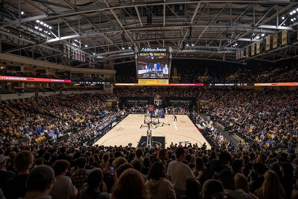 Inside a crowded basketball arena with fans seated around the court, which has a game in progress, and a large screen hanging above showing the game.