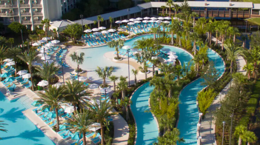 Aerial view of a resort swimming pool area with lounge chairs, umbrellas, and a lazy river surrounded by palm trees and greenery.