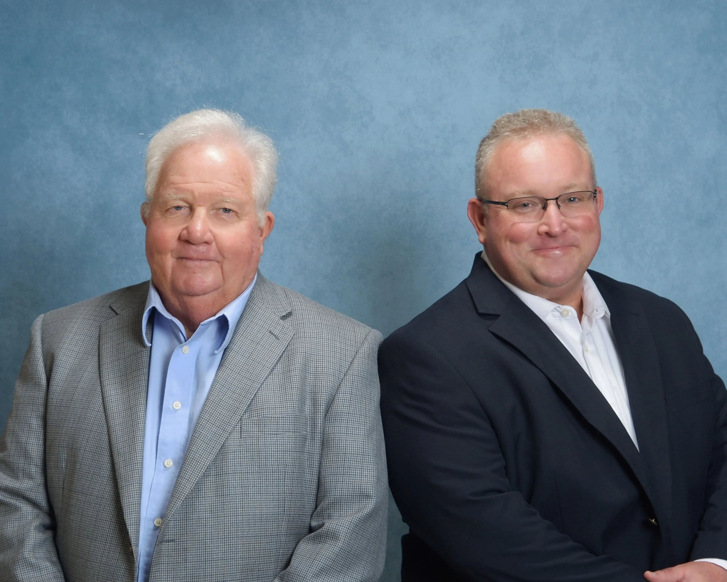 Two men in formal attire sitting against a blue background, both smiling at the camera.