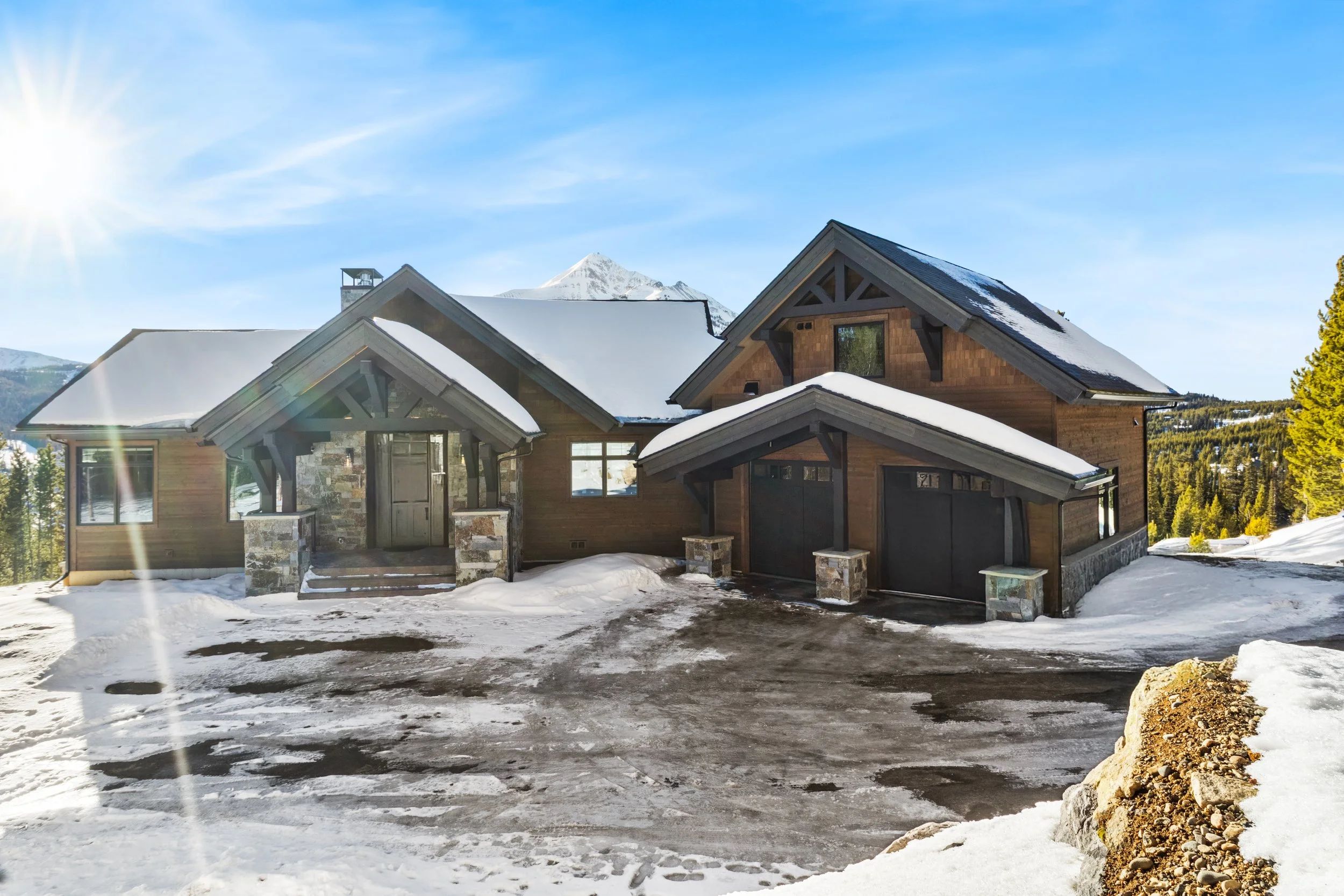 A snowy mountain landscape with a house in the foreground, mountain peaks in the background, and clear blue sky with the sun shining.