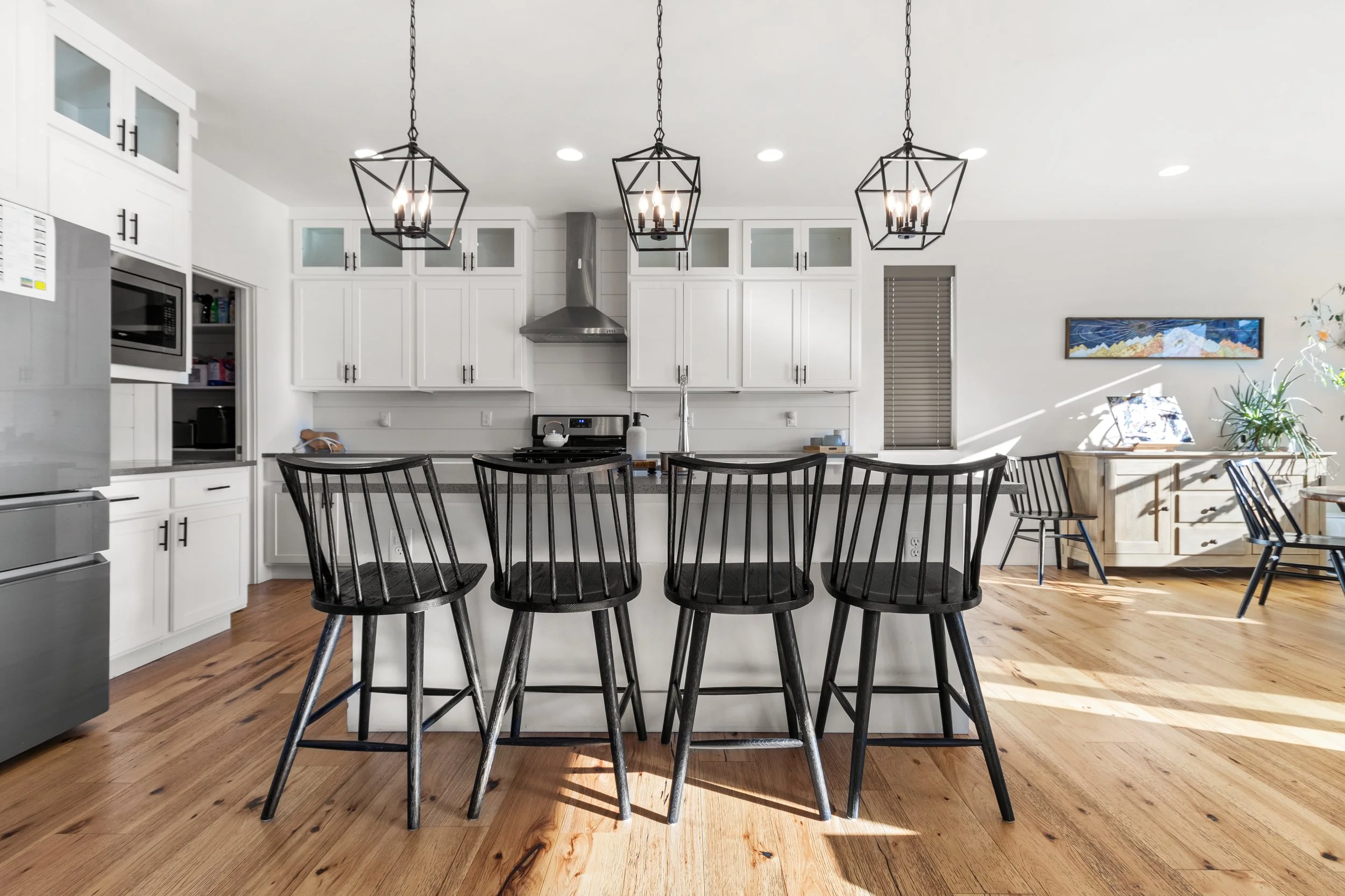 Kitchen with white cabinets, black lighting fixtures, black chairs at a kitchen island, and hardwood flooring.