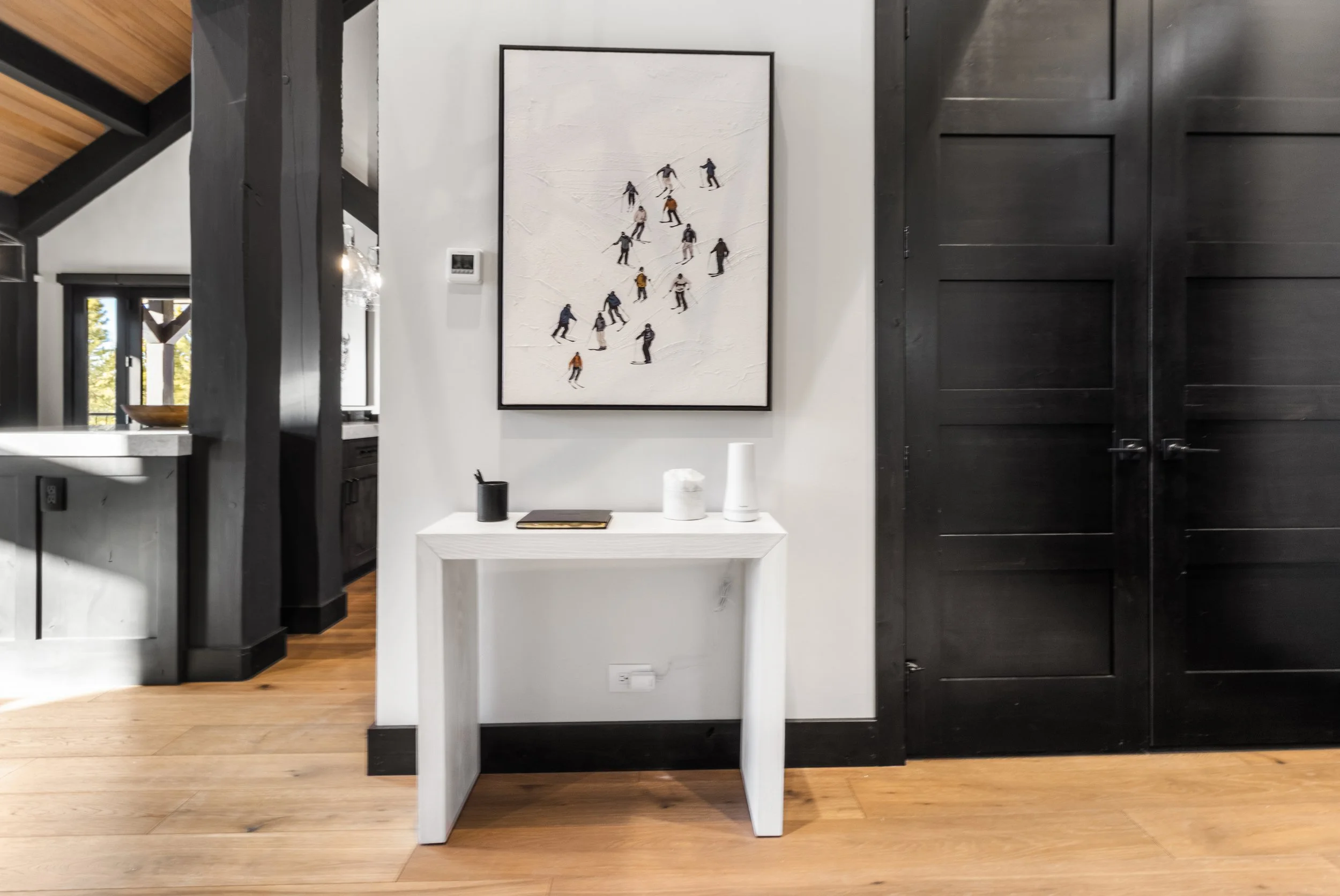 Interior view of a modern home with black and white decor, featuring a white table with vases, black candle holder, and a book, a wall art of skiers, black door, and hardwood floors.
