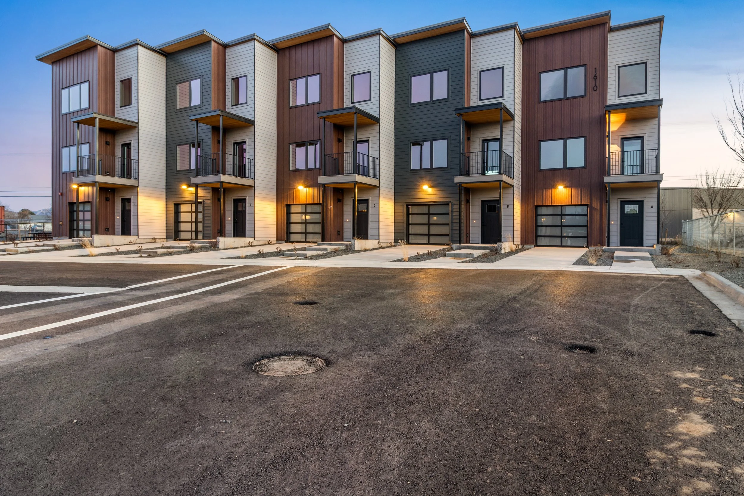 A modern multi-story residential building with a colorful exterior made of brown, beige, and dark gray panels, featuring small balconies with black metal railings, large windows, and garage doors on the ground level. The building is illuminated by exterior lights, and there is an empty parking lot with marked spaces in the foreground.