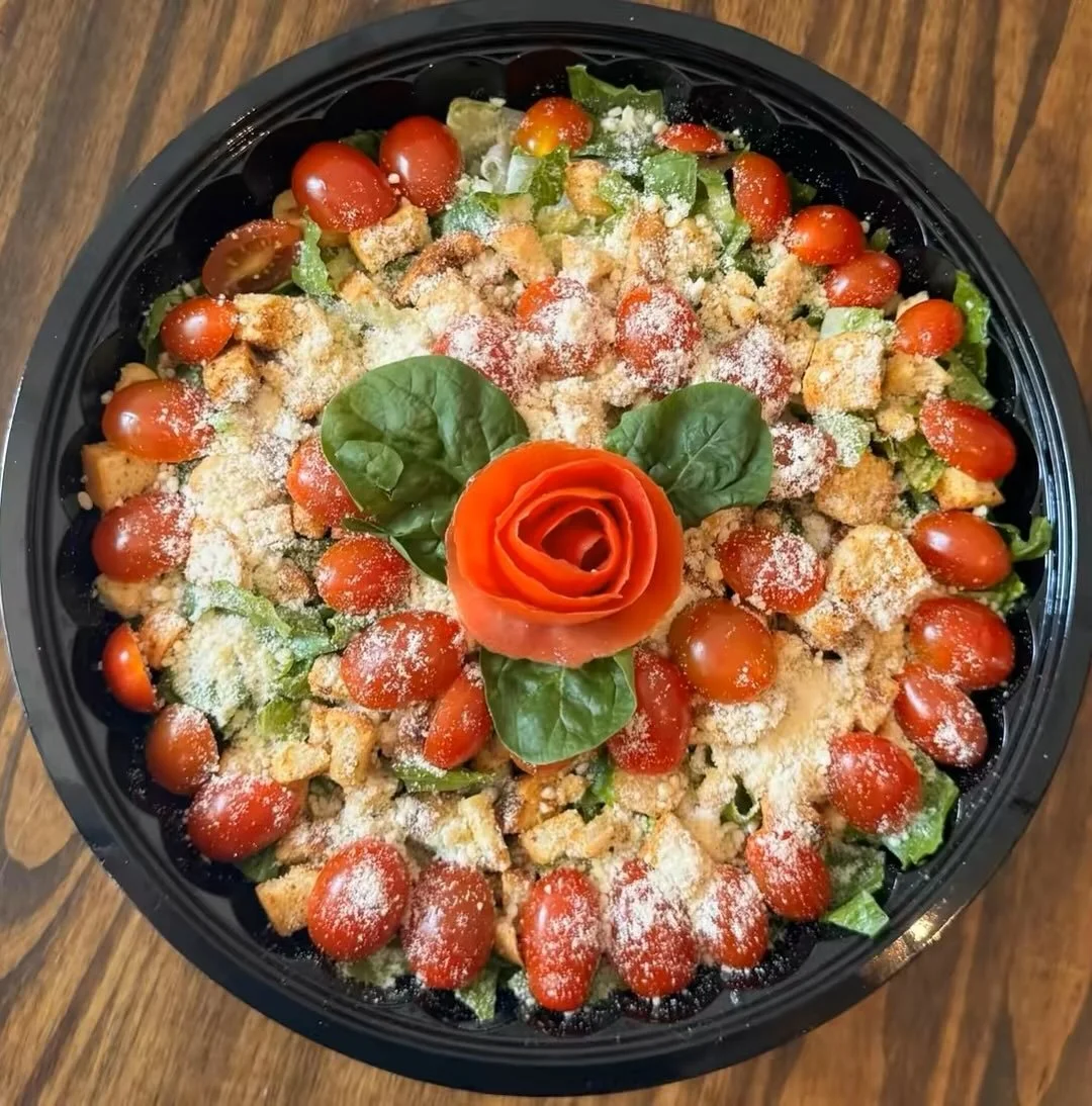 A circular salad with cherry tomatoes, lettuce, croutons, and grated cheese, topped with a decorative tomato rose and basil leaves, on a wooden surface.