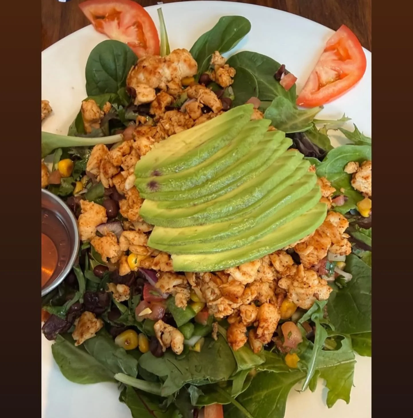 A salad with leafy greens, sliced avocado, grilled chicken, cherry tomatoes, corn, black beans, onions, and a small container of dressing on the side.