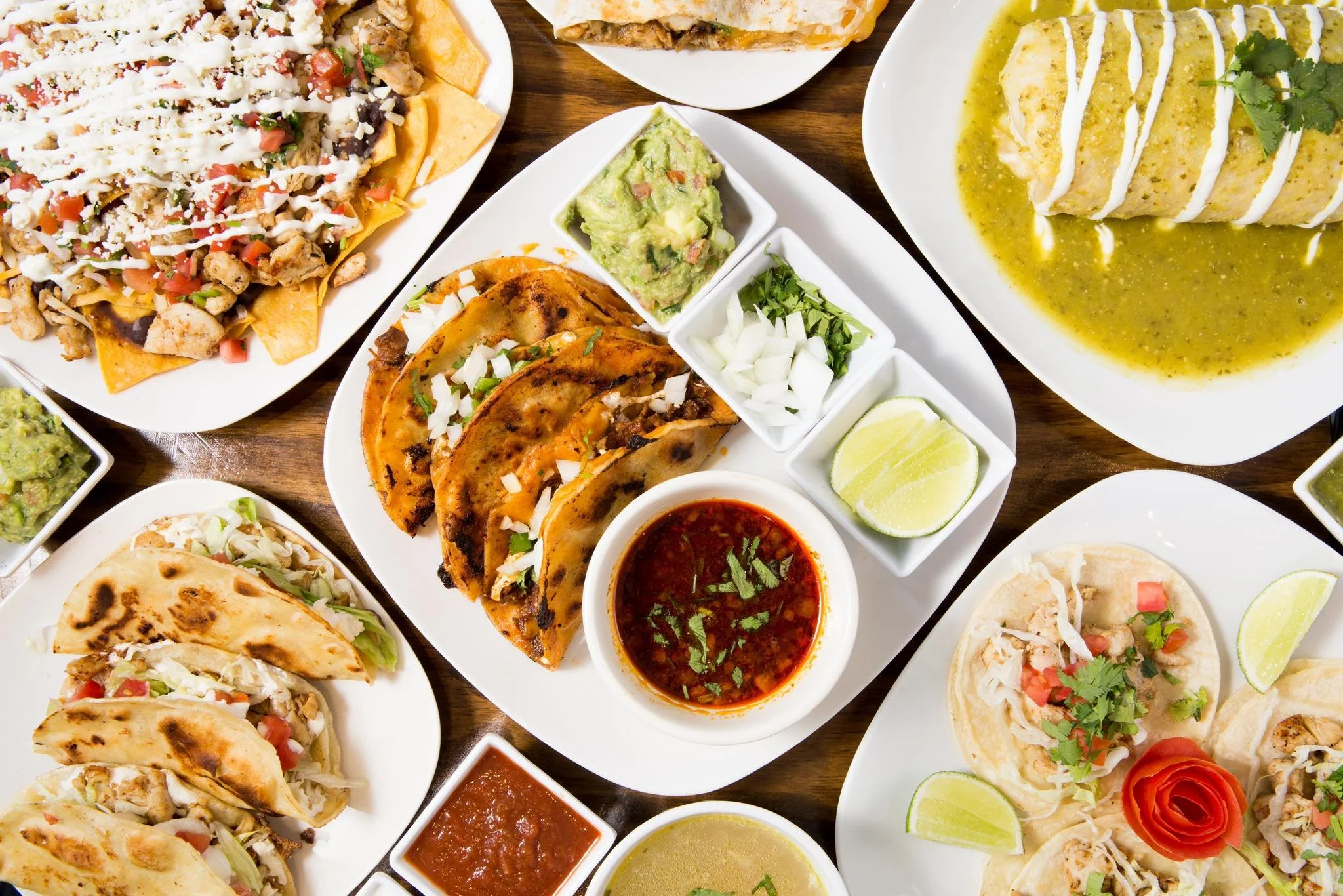 Assorted Mexican dishes including tacos, nachos, enchiladas, guacamole, pico de gallo, lime wedges, and salsa on white plates on a wooden table.