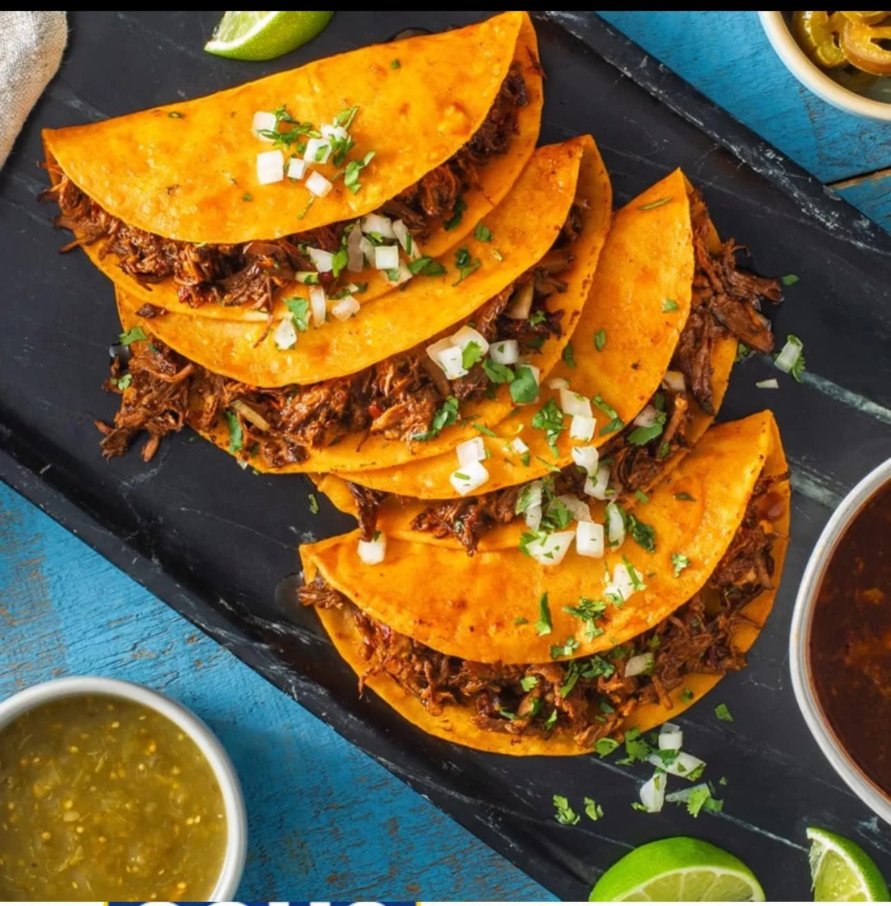 Six Mexican tacos filled with shredded meat, topped with chopped onions and cilantro, served on a black platter with lime wedges, green salsa, and dark sauce.