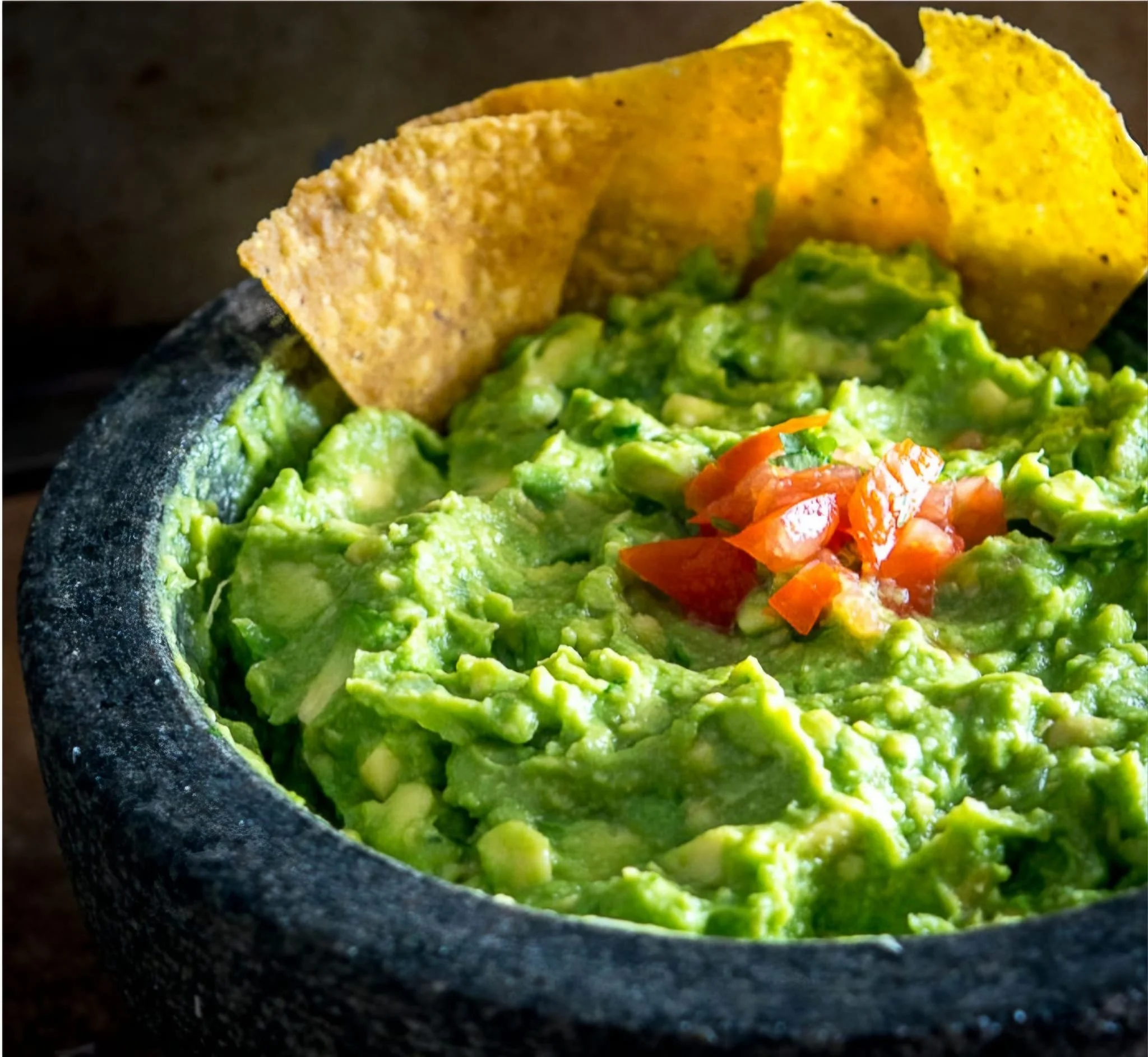 A black stone bowl filled with chunky guacamole topped with chopped tomatoes and cilantro, with tortilla chips in the background.