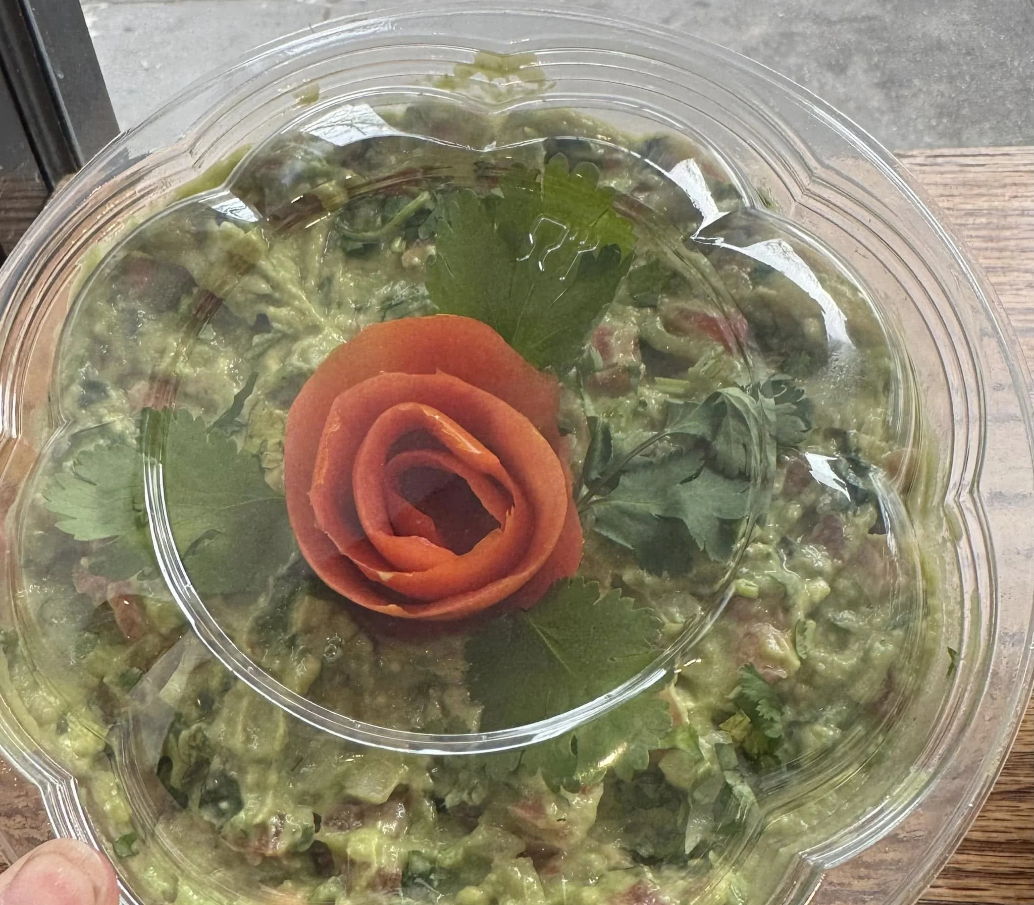 A plastic takeout container of guacamole topped with a decorative tomato flower and cilantro leaves, sitting on a wooden table.