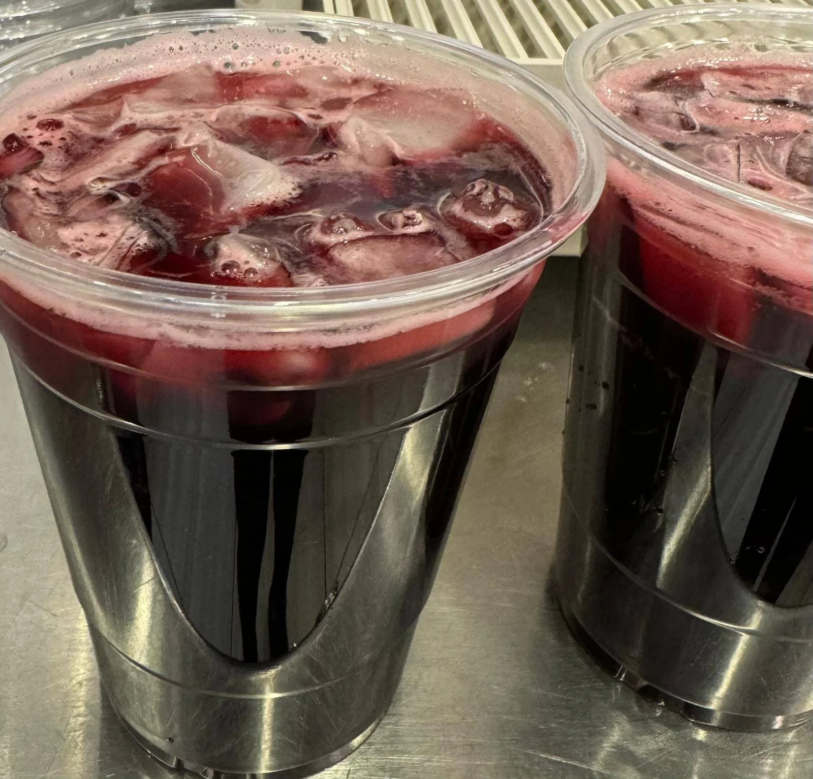 Two clear plastic tumblers filled with a dark red beverage, ice cubes, and floating cranberries, placed on a metallic surface.