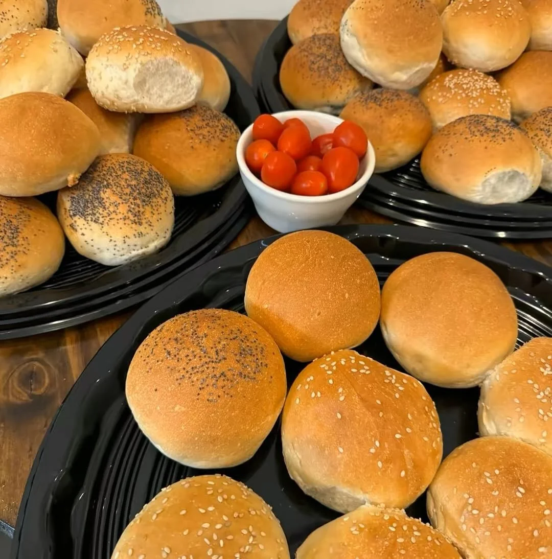 A variety of hamburger buns with different toppings, arranged on black plastic trays, with a small bowl of cherry tomatoes in the center.