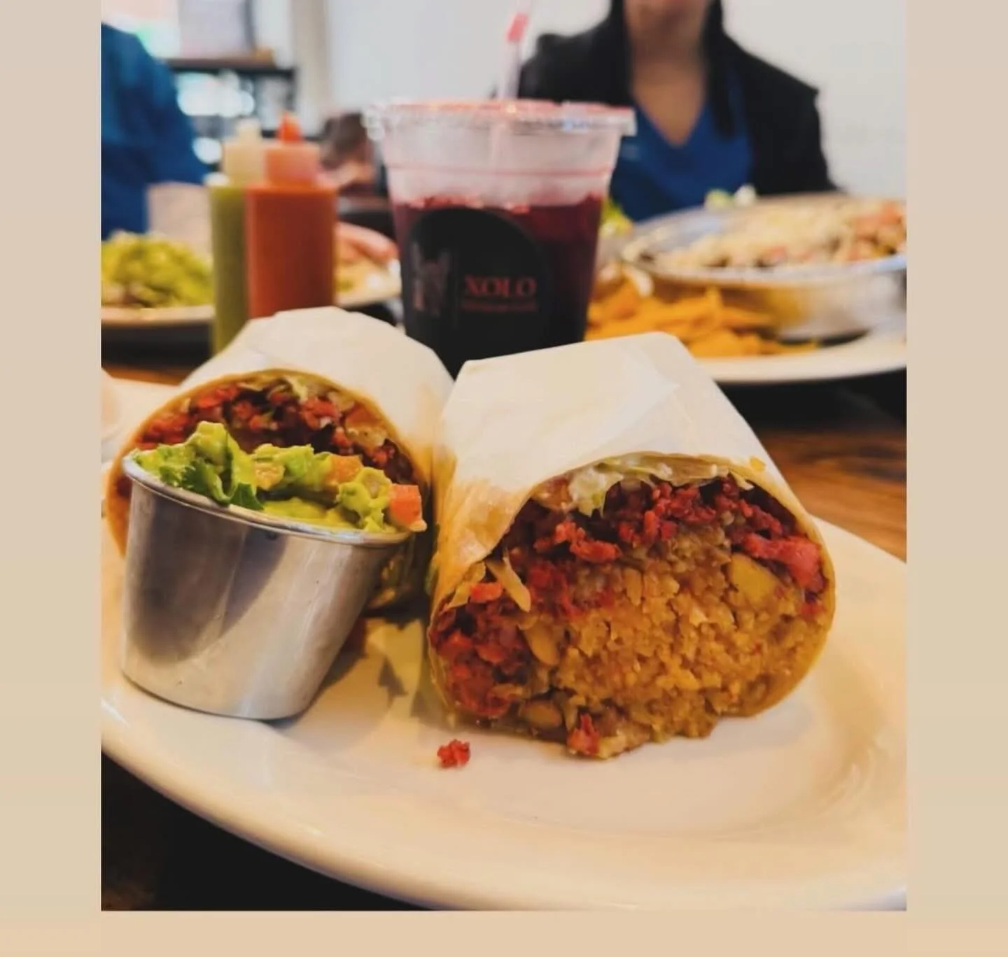 A plate with two burritos, one with guacamole in a metal cup, on a wooden table at a restaurant, with a glass of dark soda and other dishes in the background.