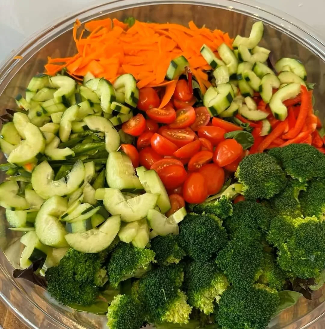 A glass bowl filled with chopped vegetables including cherry tomatoes, broccoli, sliced cucumbers, shredded carrots, and sliced green bell peppers.