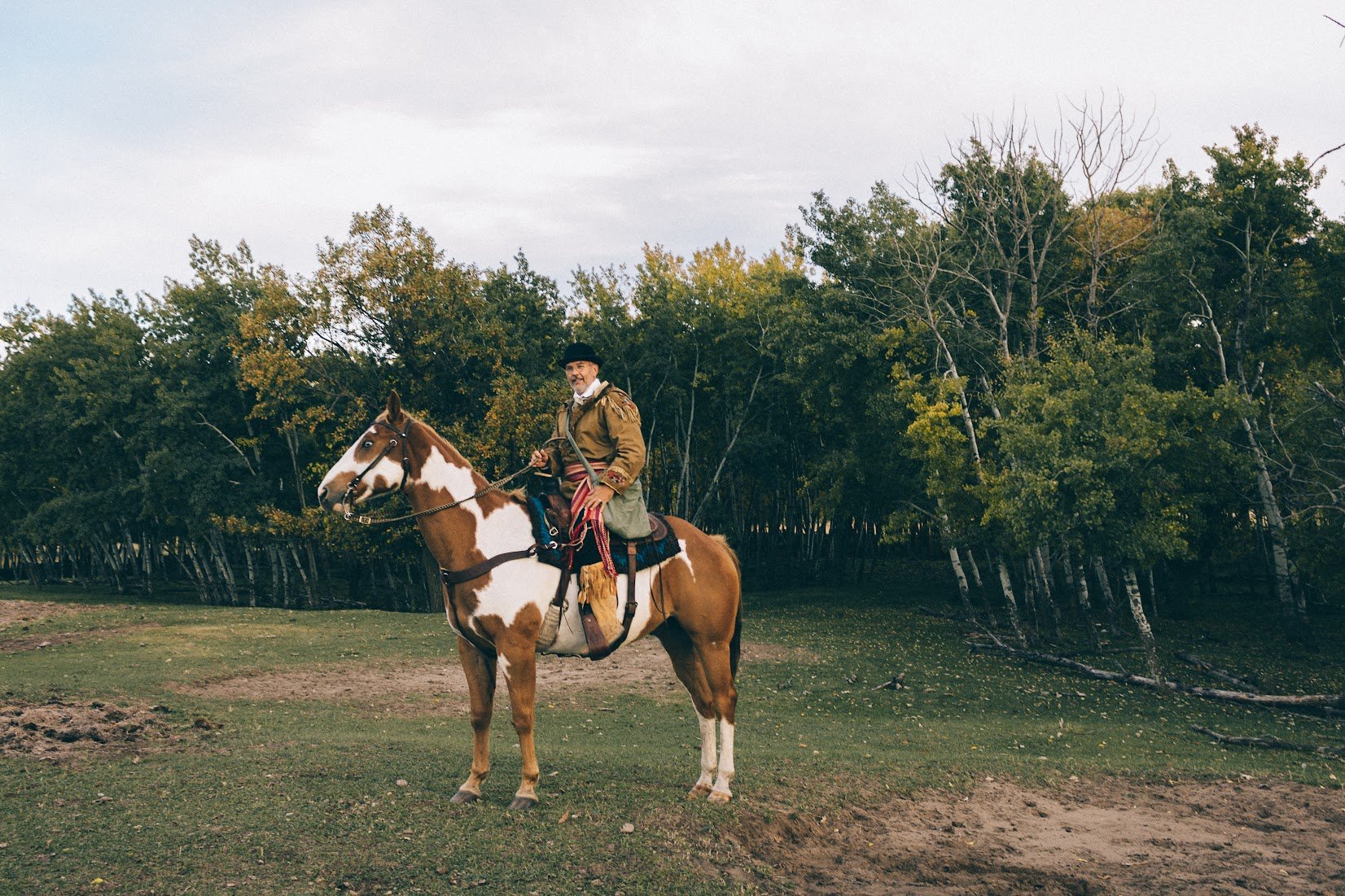 Author Denis Fortier dressed in historical clothing, riding a paint horse in an outdoor setting with trees in the background.
