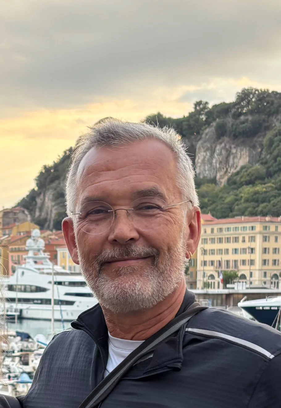 Author Denis Fortier, wearing glasses and a black jacket, standing near a marina with yachts, colorful buildings, and a hillside in the background during sunset.