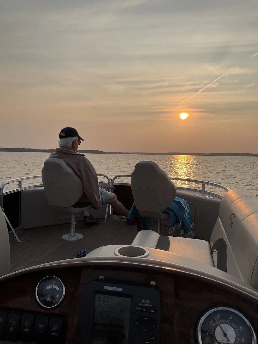 A person sitting on a boat during sunset, looking out over calm water with a distant shoreline, in the boat's cockpit area with navigational equipment visible.
