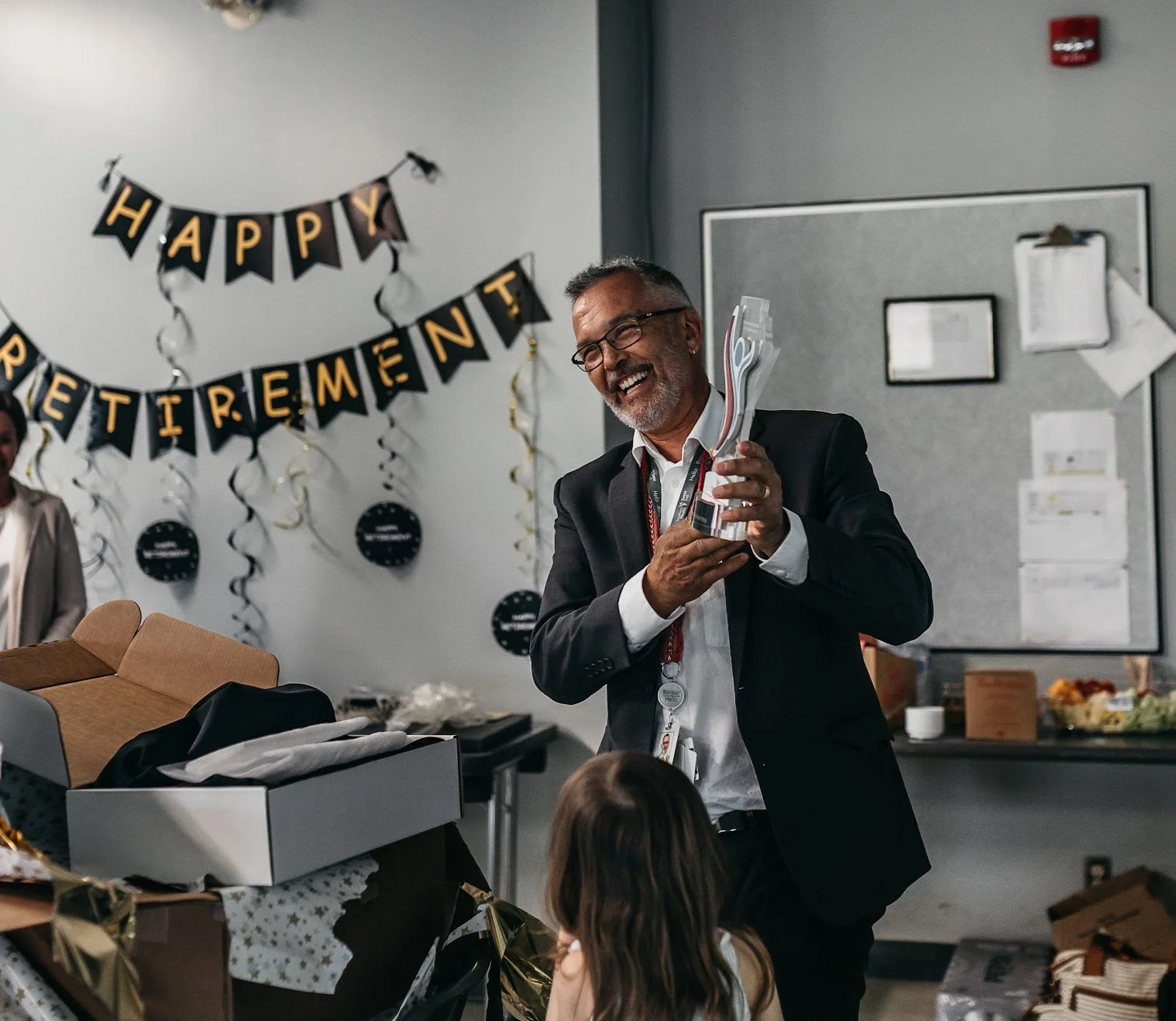 Author Denis Fortier in a suit holding a trophy and smiling at a celebration party with a 'Happy Retirement' banner in the background.