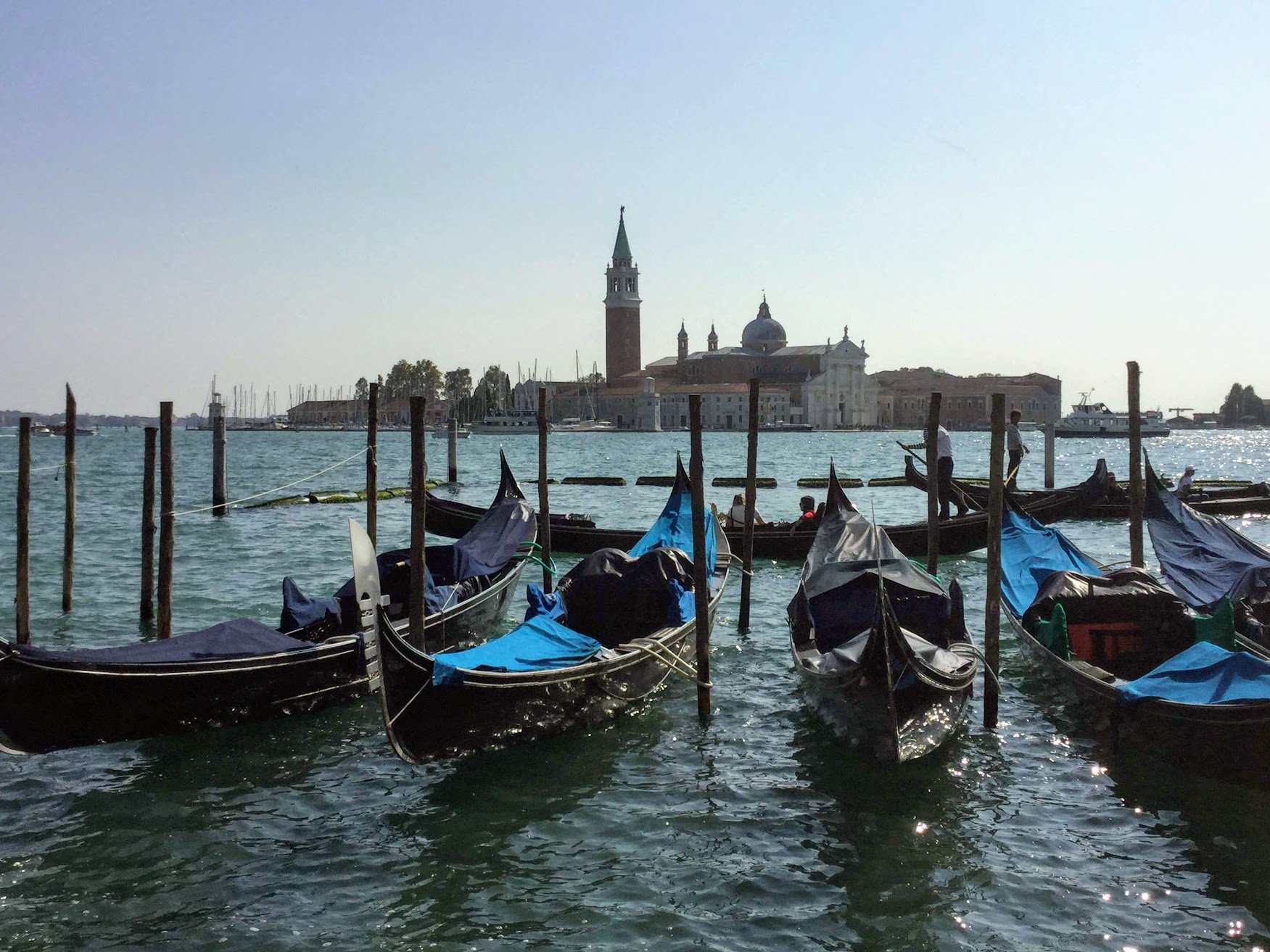 Gondolas docked along a waterfront with a historic church and bell tower in the background, and boats sailing on the water.