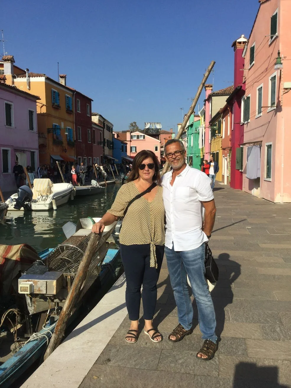 Author Denis Fortier and his wife standing on a canal-side walkway in colorful buildings in Burano, Italy, with boats docked in the canal behind them.