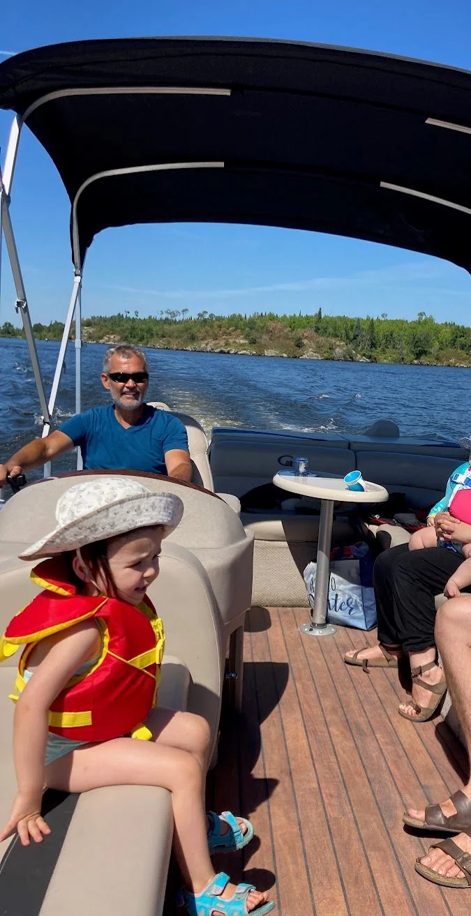 A family enjoying a boat ride on a sunny day with clear skies, sitting on a boat with a canopy, water and trees in the background, a young girl in a life jacket and hat, an older man smiling, and partially visible adult women.