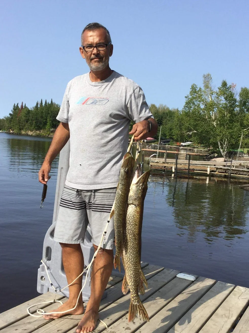  Author Denis Fortier standing on a wooden dock holding two large fish he caught, with trees and water in the background on a clear day.