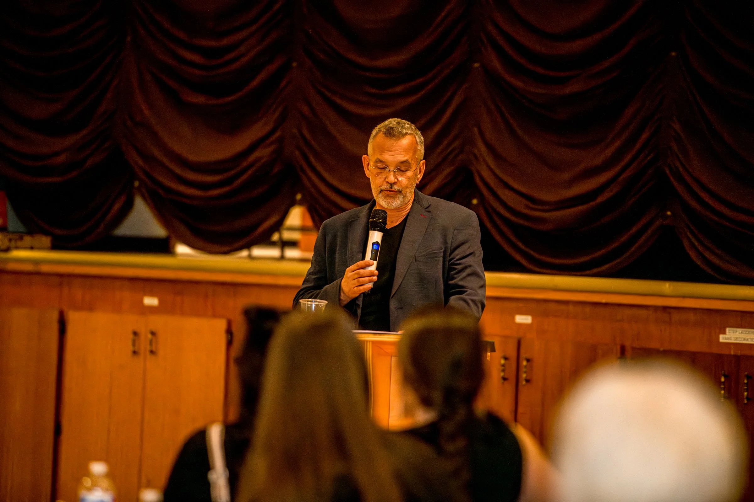 Author Denis Fortier speaking into a microphone at a podium during an event or presentation, with a large closed red velvet curtain behind him, in a room with an audience.
