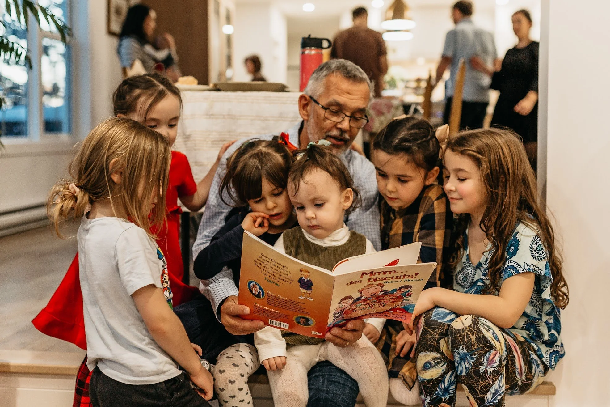 Author Denis Fortier reading a children's book titled 'Mmm... des Biscuits' surrounded by six grand children, all focused on the book, in a warmly lit indoor setting with other people in the background.