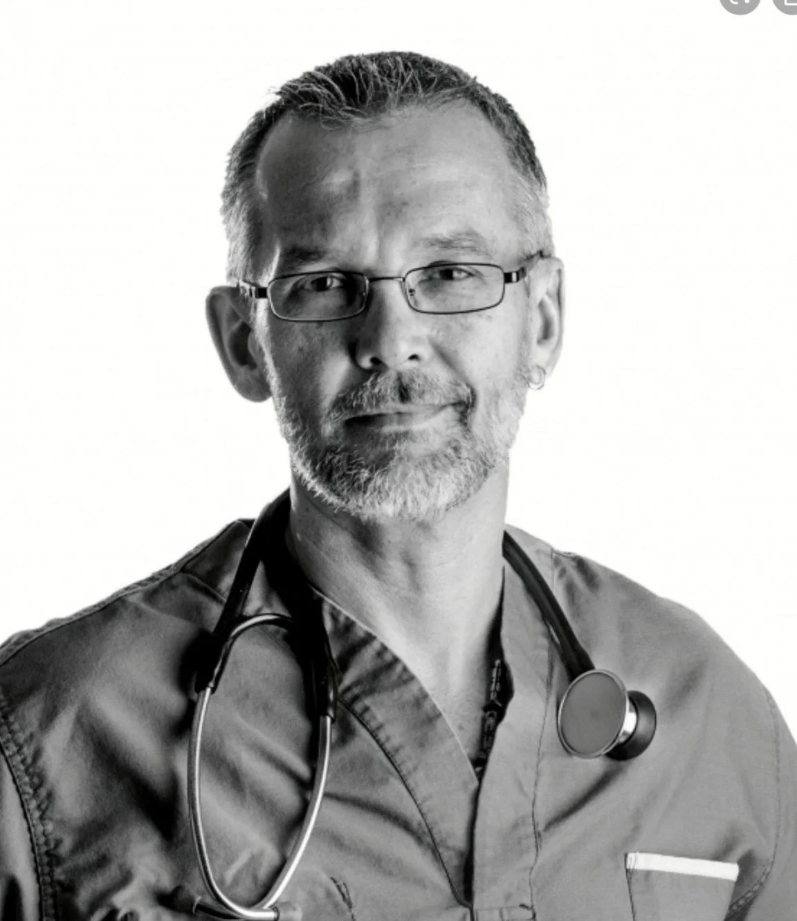 Black and white portrait of a male healthcare professional with glasses, gray hair, a beard, and a stethoscope around his neck, wearing scrubs.