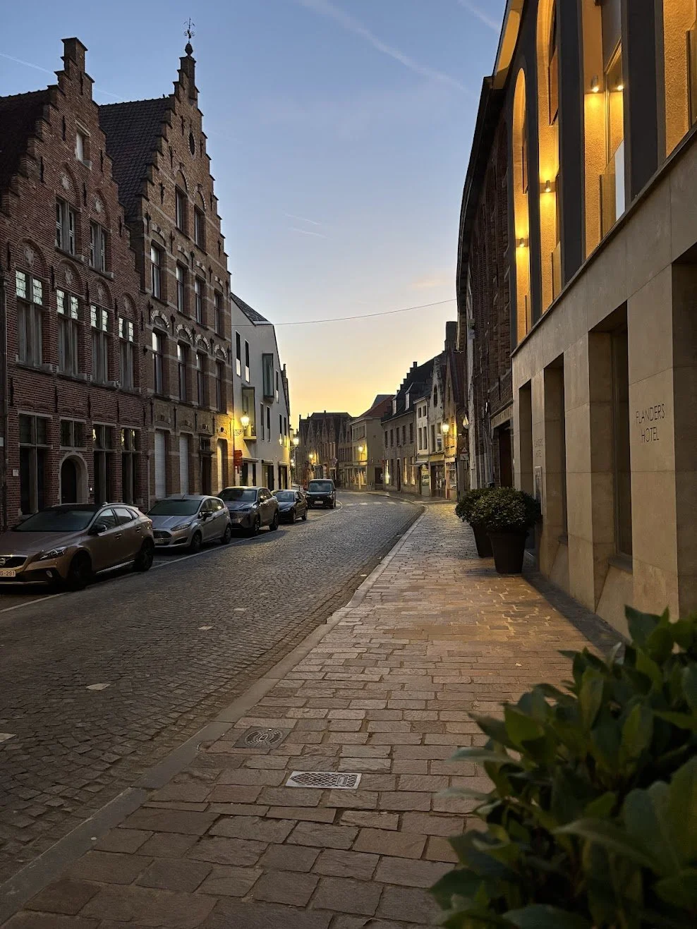 A quiet city street during sunset with parked cars on one side, historic brick buildings on the left, modern buildings with warm lighting on the right, and a cobblestone sidewalk and street.