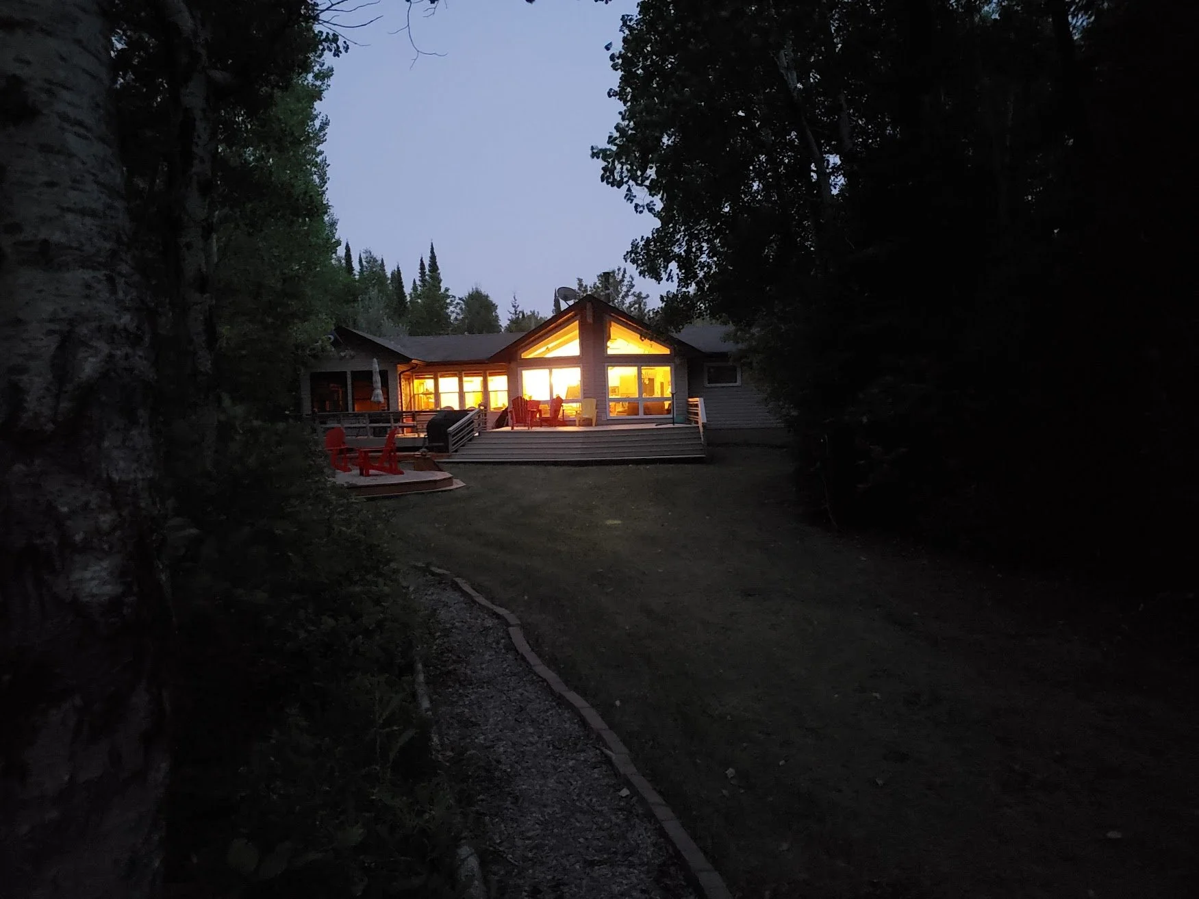 A cottage with large illuminated windows on a deck, surrounded by trees at dusk or night.