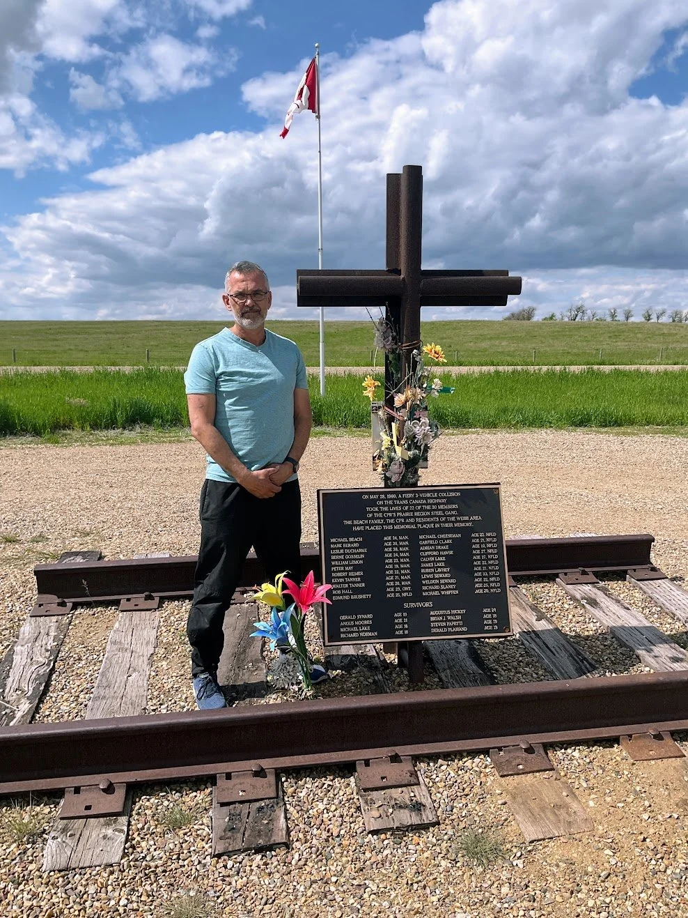 Author Denis Fortier standing on a memorial for the CP Railway's Prairie Steel Gang marked by a large black cross and flowers, next to a railroad track. In the background, an open field, a flagpole with a Canadian flag, and a partly cloudy sky.