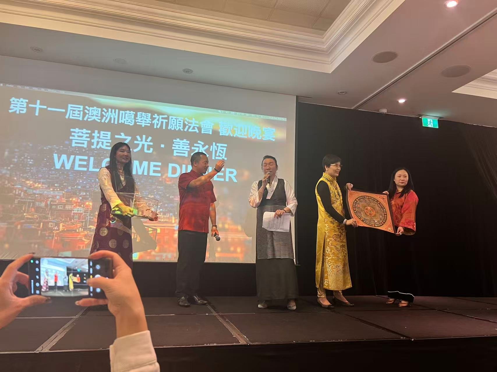 A group of five individuals on stage during an event, with a large screen behind them displaying Chinese characters and the words 'WELCOME DINNER'. Three women and two men, some in traditional attire, are engaged in an award or gift exchange, with on