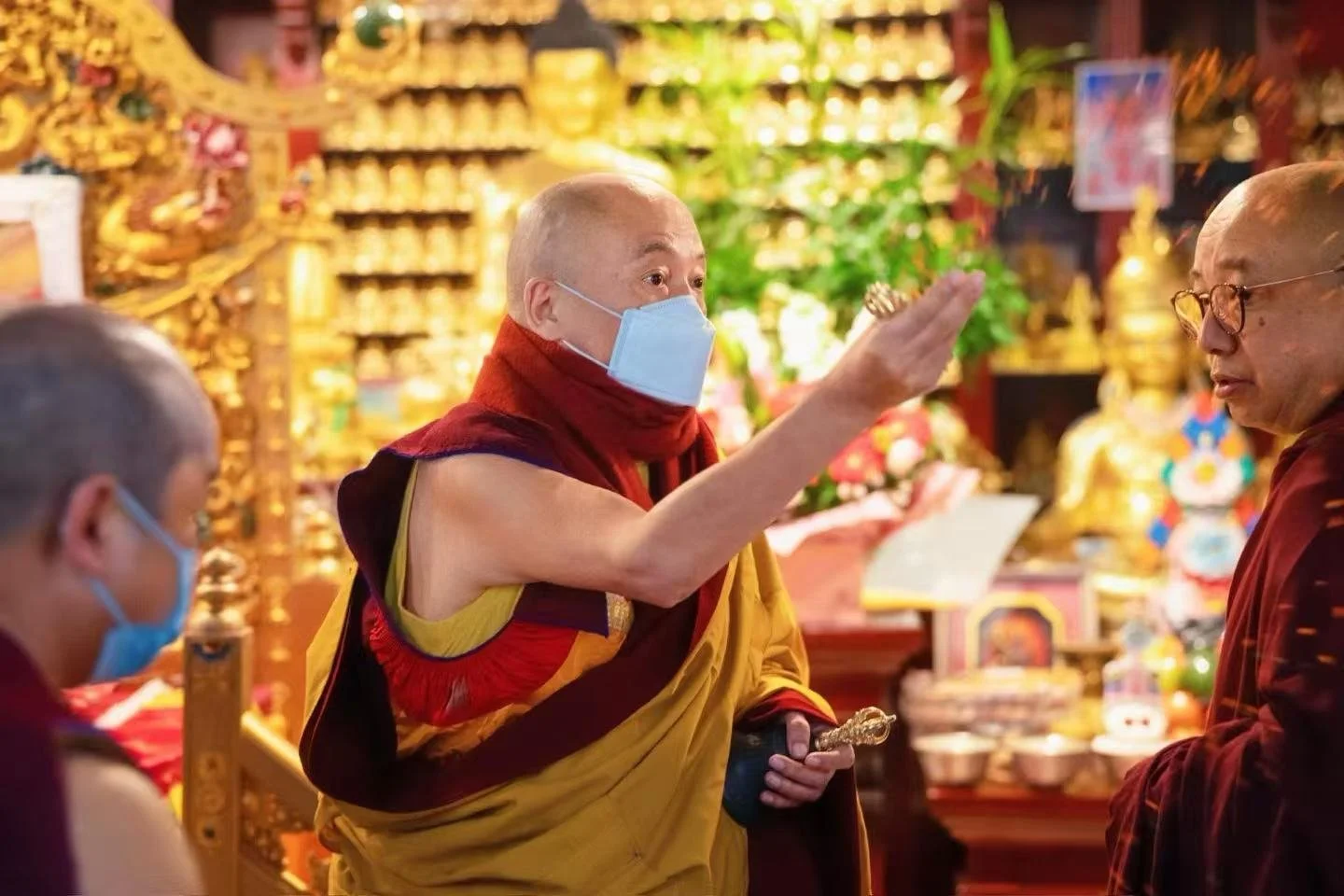 Glaytsab Rimpoche. Buddhist monks wearing face masks engaged in a discussion inside a temple with golden statues and colorful decorations in the background. Australia Kamtsang Dharma Center