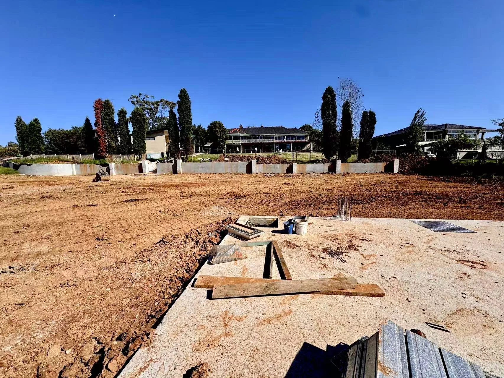 Construction site with dirt ground, construction materials, and partially poured concrete foundation, with residential houses and trees in the background under a clear blue sky.
