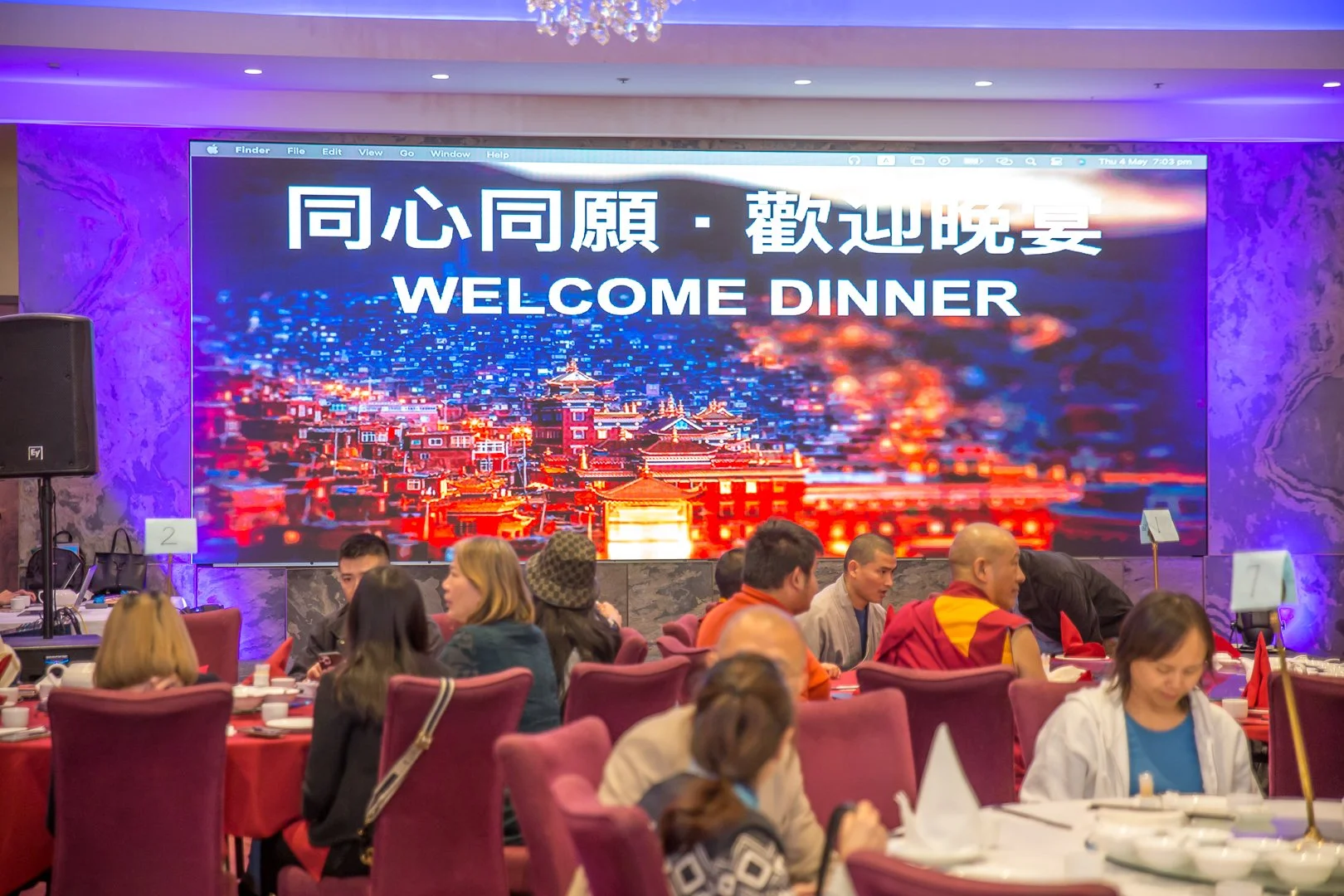 Large digital screen with a cityscape background and text in Chinese characters with 'Welcome Dinner' in English. People seated at round tables with red tablecloths are dining in a banquet hall.