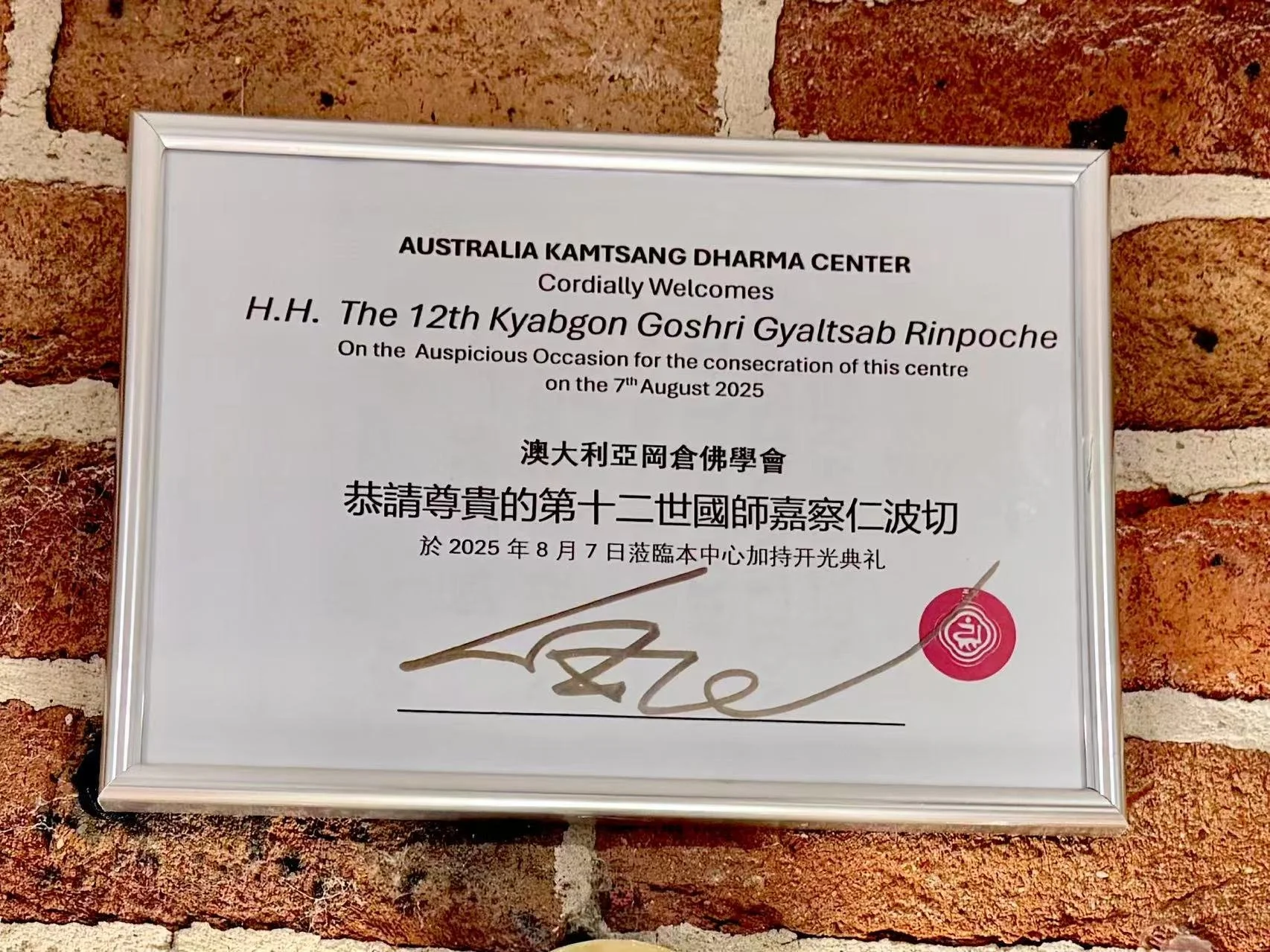 A framed certificate on a brick wall. The certificate is from Australia KAMTSANG DHARMA CENTER welcoming H.H. The 12th Kyabgon Goshri Gyaltsab Rinpoche for a consecration event on August 7, 2025, with signatures and a red official stamp.