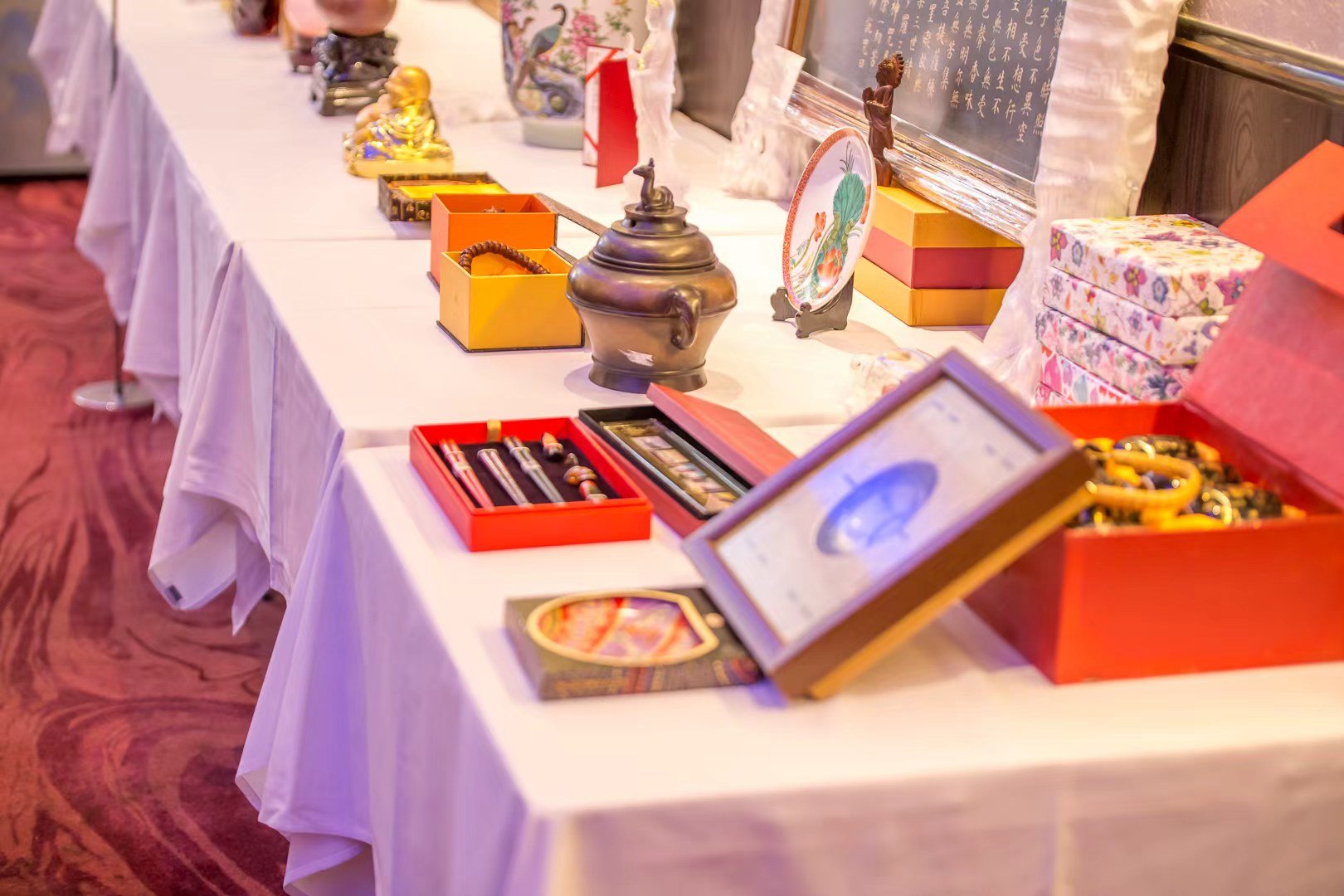 A display table with various decorative and collectible items including boxes, jewelry, a teapot, and figurines, arranged along a white cloth tablecloth at an indoor event.