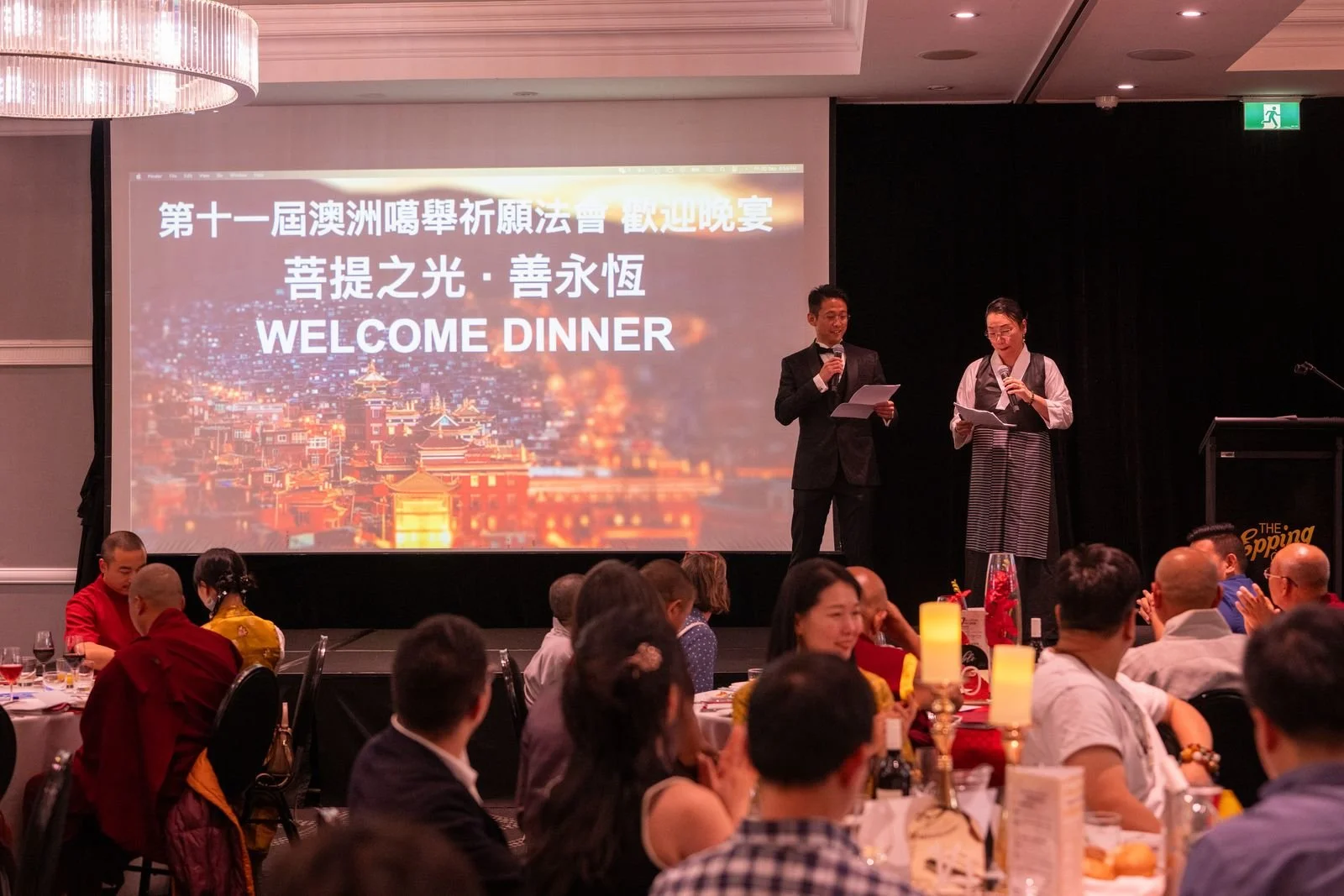 A large screen on stage displays Chinese and English text welcoming guests to a dinner event. Two people stand on stage, speaking into microphones. The room is filled with seated attendees, some clapping, with tables decorated with candles and flower
