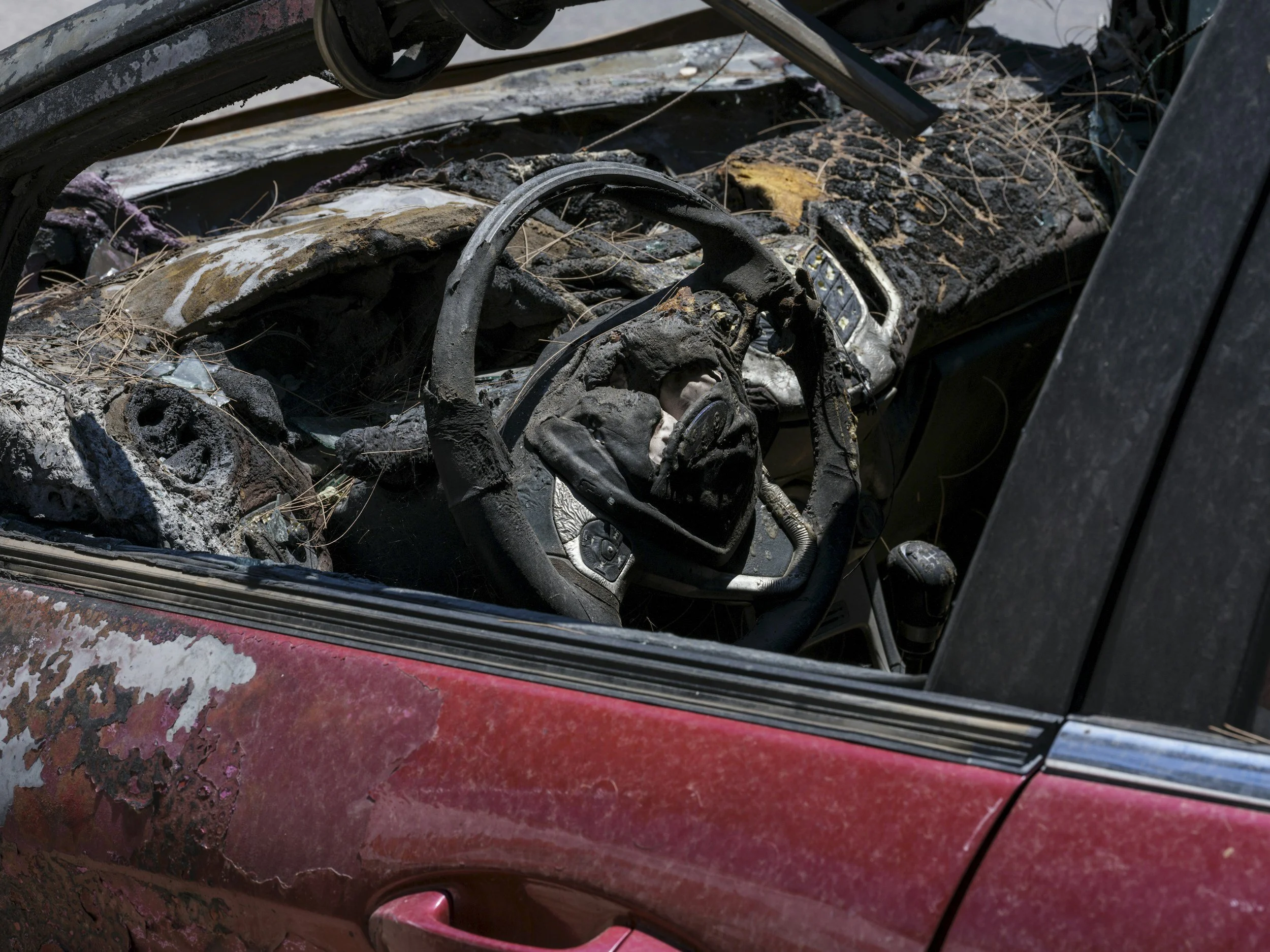 The image shows the interior of a severely burned car with a melted dashboard, charred steering wheel, and debris scattered inside. The exterior features peeling or damaged paint.