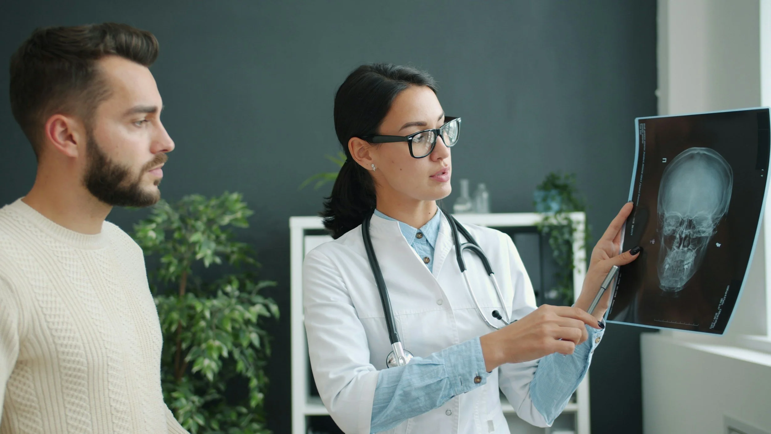 Female doctor holding an X-ray image of a skull, discussing it with a male patient in a medical office.