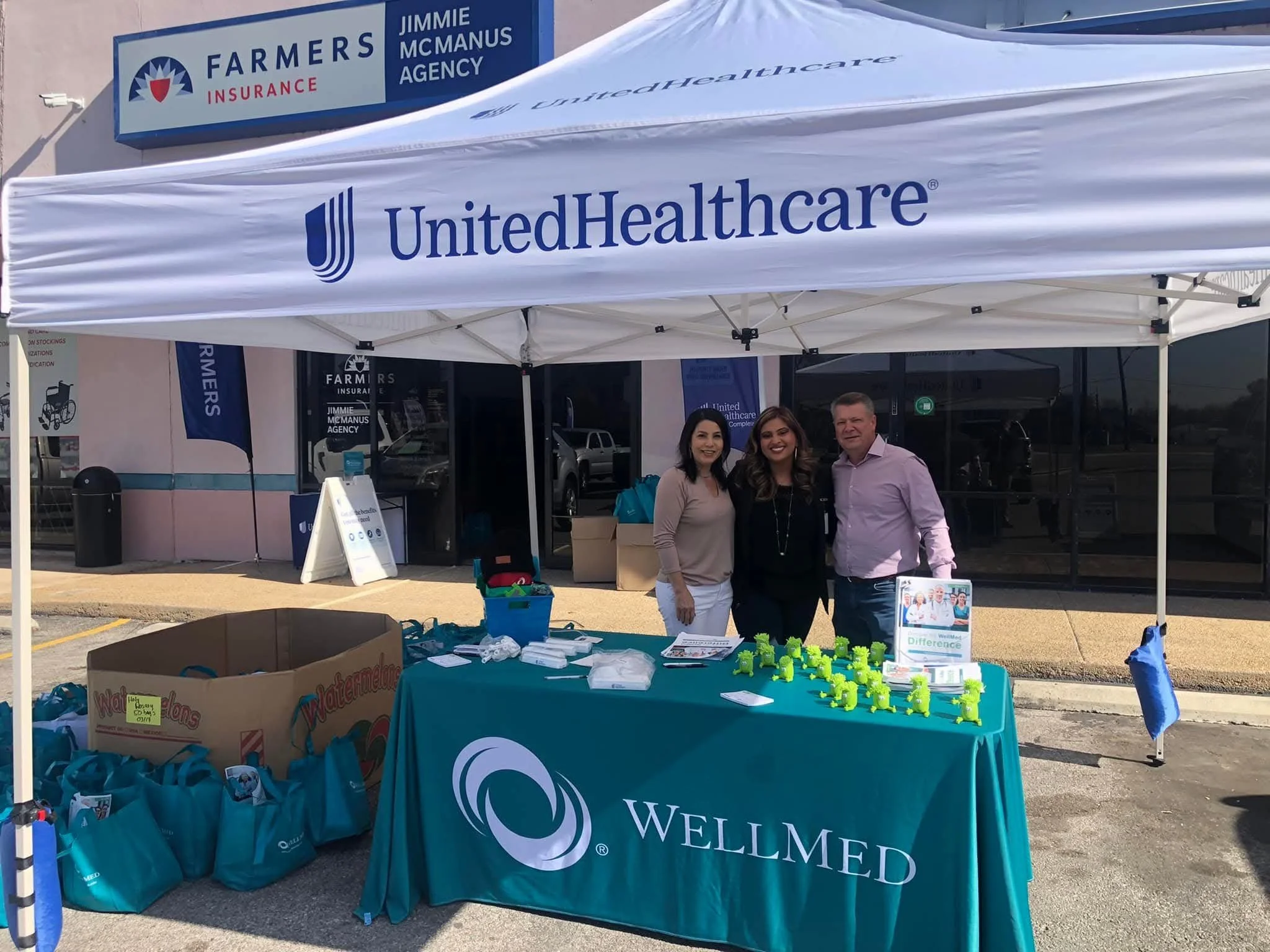 A group of three people standing under a white tent with  a0United Healthcare a0 and Welmed branding. The tent is set up outside a building with a Farmer a0 Insurance sign. In front of the people, there is a table with various items including green s