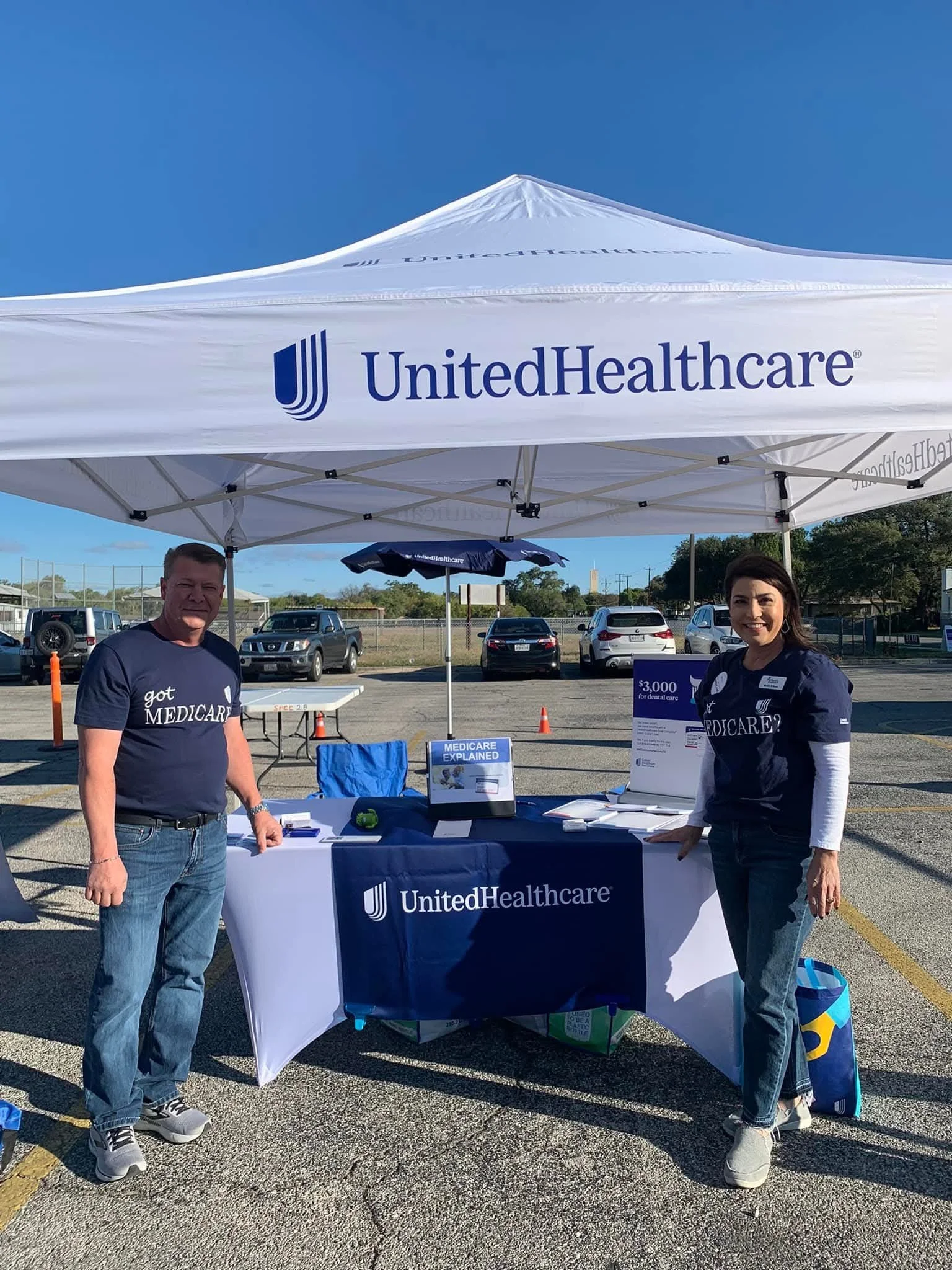 Two people standing under a UnitedHealthcare canopy at an outdoor health event, with tables and informational signage about Medicare and dental care, in a parking lot.