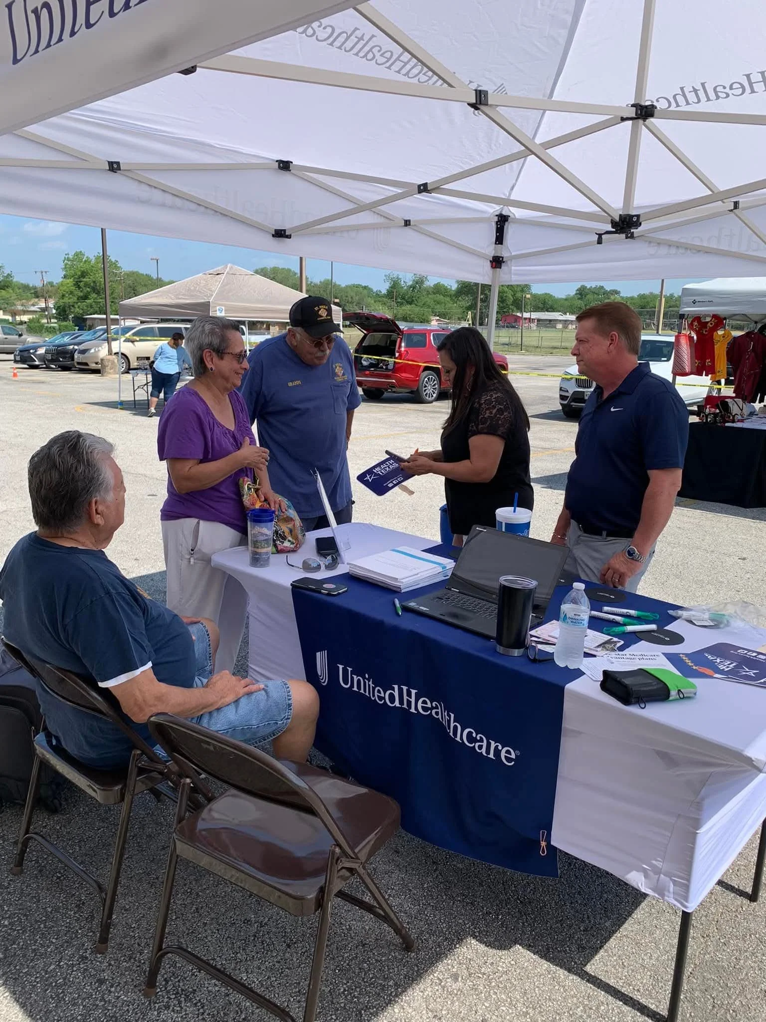 People gathered under a white tent at an outdoor event with a table displaying UnitedHealthcare materials, and a man and woman talking to a woman holding a phone. Vehicles are parked nearby.